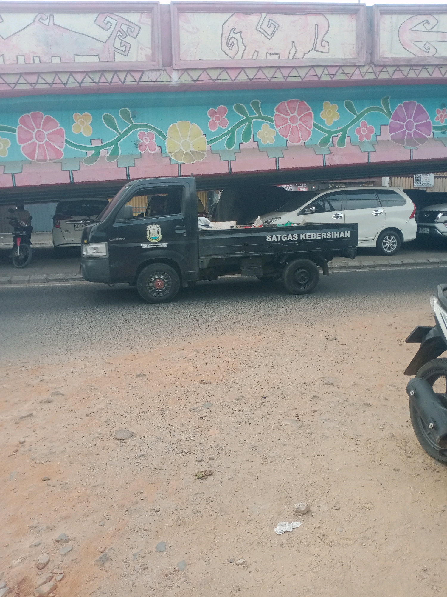 The image depicts a black Suzuki Carry pickup truck parked on a dirt shoulder next to a paved road. The truck has the words "SATGAS KEBERSIHAN" (Sanitation Task Force) written in white on its side. A white minivan and a motorcycle are also visible in the background, along with a section of an overhead structure decorated with colorful floral and abstract motifs. The scene appears to be in an outdoor urban or semi-urban environment. The dirt shoulder is scattered with small stones and debris, and a piece of white crumpled material is visible on the ground near the lower right corner. The overall lighting suggests it is daytime.