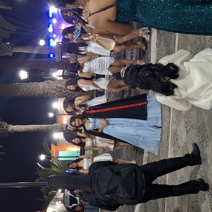 A group of young women in formal attire line a stone path at night, likely for a celebration. Dresses range from white and light blue to emerald green. A man with a guitar case stands at the front. Palm trees and streetlights line the path, leading to a structure illuminated with colored lights. The scene suggests a quinceañera or similar event.