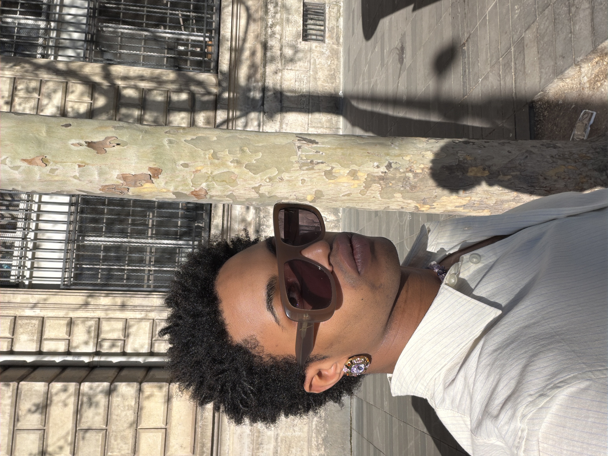 A medium shot shows a Black person with brown sunglasses, and an earring, standing near a sycamore tree in what appears to be Paris, France. The person is wearing a striped white shirt with a partially visible amethyst necklace. The tree trunk is behind them, with a light yellowish-green trunk that has peeling bark. The bark is a combination of yellow, green, brown, and off-white colors. Behind the tree, there's a building with barred windows and a weathered facade, indicating an urban environment. A sidewalk is visible on the right edge of the frame, providing a sense of the surroundings. The image composition includes elements of nature and architecture, placing the person in an outdoor urban setting. A shadow of a person is visible on the sidewalk.