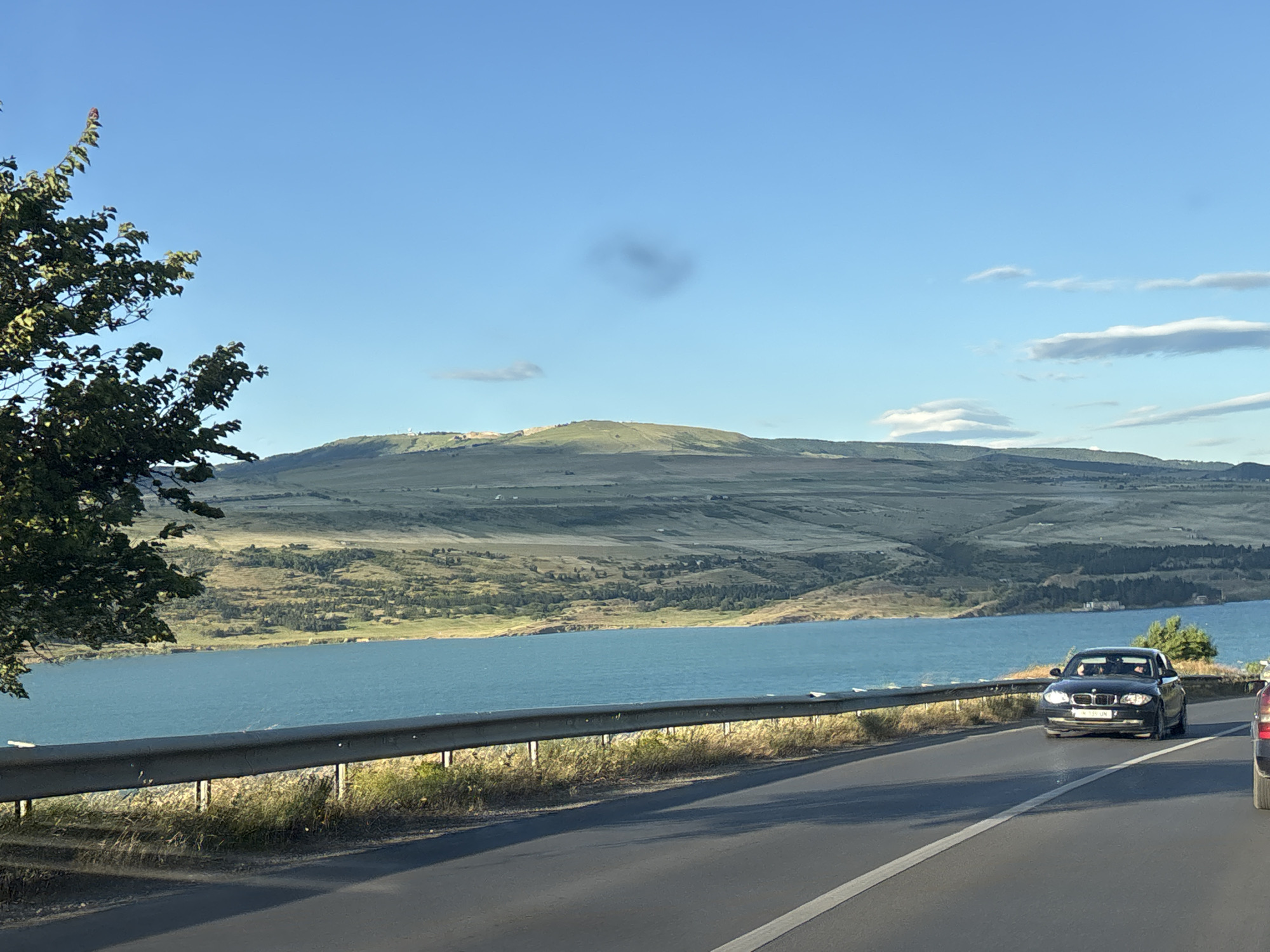 The image captures a scenic view from a road overlooking a large body of turquoise water, likely a reservoir or lake, with rolling hills in the background. A black BMW sedan is driving on the road in the foreground, with at least two individuals visible inside. The road is bordered by a metal guardrail and sparse vegetation. To the left of the frame, a leafy green tree extends into the shot. The sky is clear blue with a few wispy clouds. The scene appears to be set during daylight, likely late afternoon or early evening, given the quality of light and shadows. This view is consistent with locations found along the road to the Tbilisi Sea, a reservoir located on the northeastern outskirts of Tbilisi, Georgia.