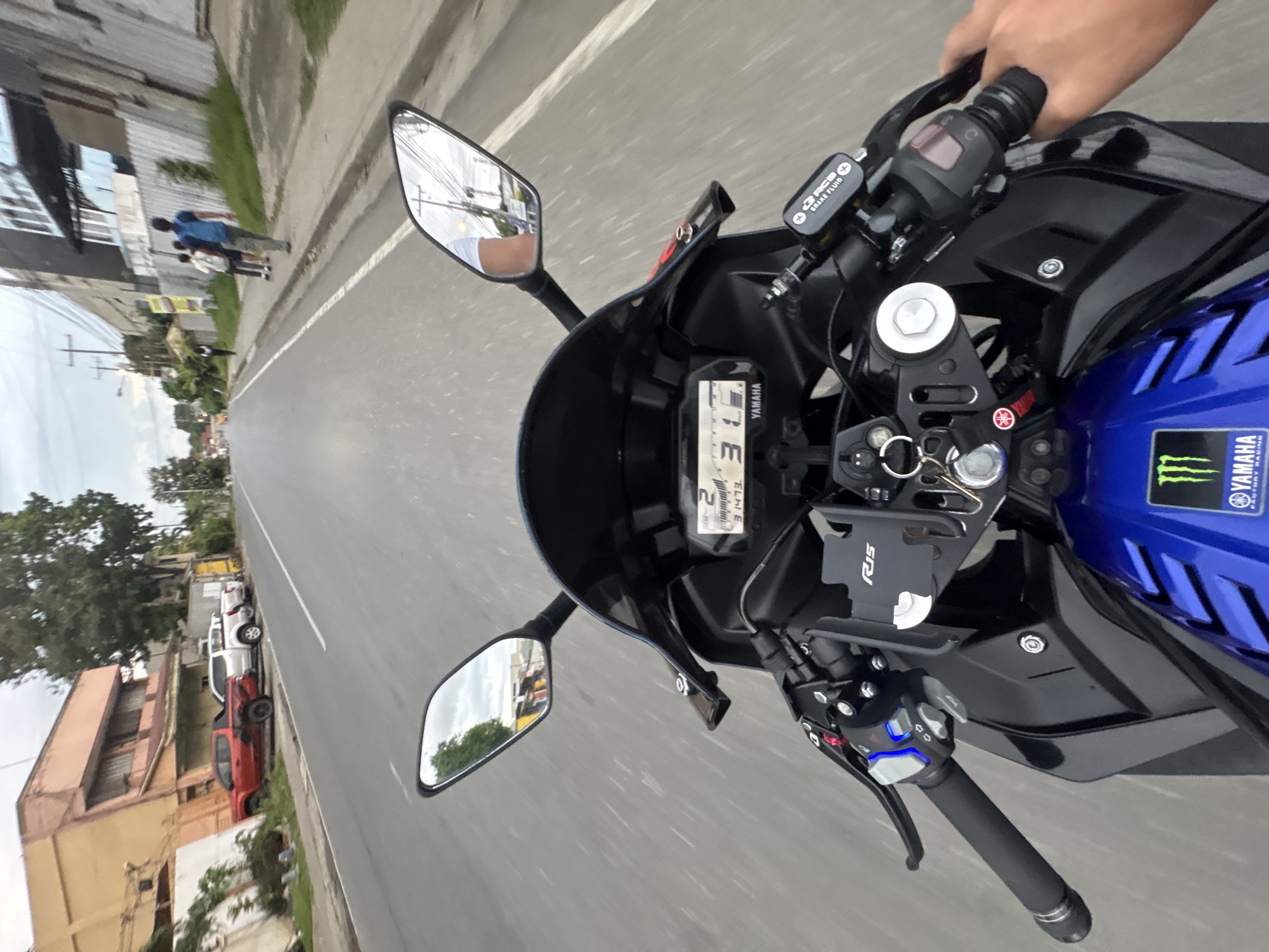 The image is a first-person perspective of someone riding a black and blue Yamaha R15 motorcycle on a paved road in Davao City, Philippines. The motorcycle's digital dashboard shows "2" in the gear indicator and "31" as the speed in kilometers per hour. The rider's right hand is on the handlebar, operating the throttle and brake.
The road appears to be a main thoroughfare with two lanes in each direction, marked by a white broken line. On the left side of the road, there is a building with an orange facade and several parked vehicles, including a red pickup truck and two white pickup trucks. Further down the road, there are trees and power lines.
On the right side of the road, there is a sidewalk with several pedestrians. One individual, wearing a blue backpack, is walking away from the camera. Behind them, two other individuals are walking in the same direction. Beyond the sidewalk, there is a building with a corrugated metal fence and some vegetation. The sky is overcast, suggesting a cloudy day. The environment suggests an urban or semi-urban setting within Davao City.