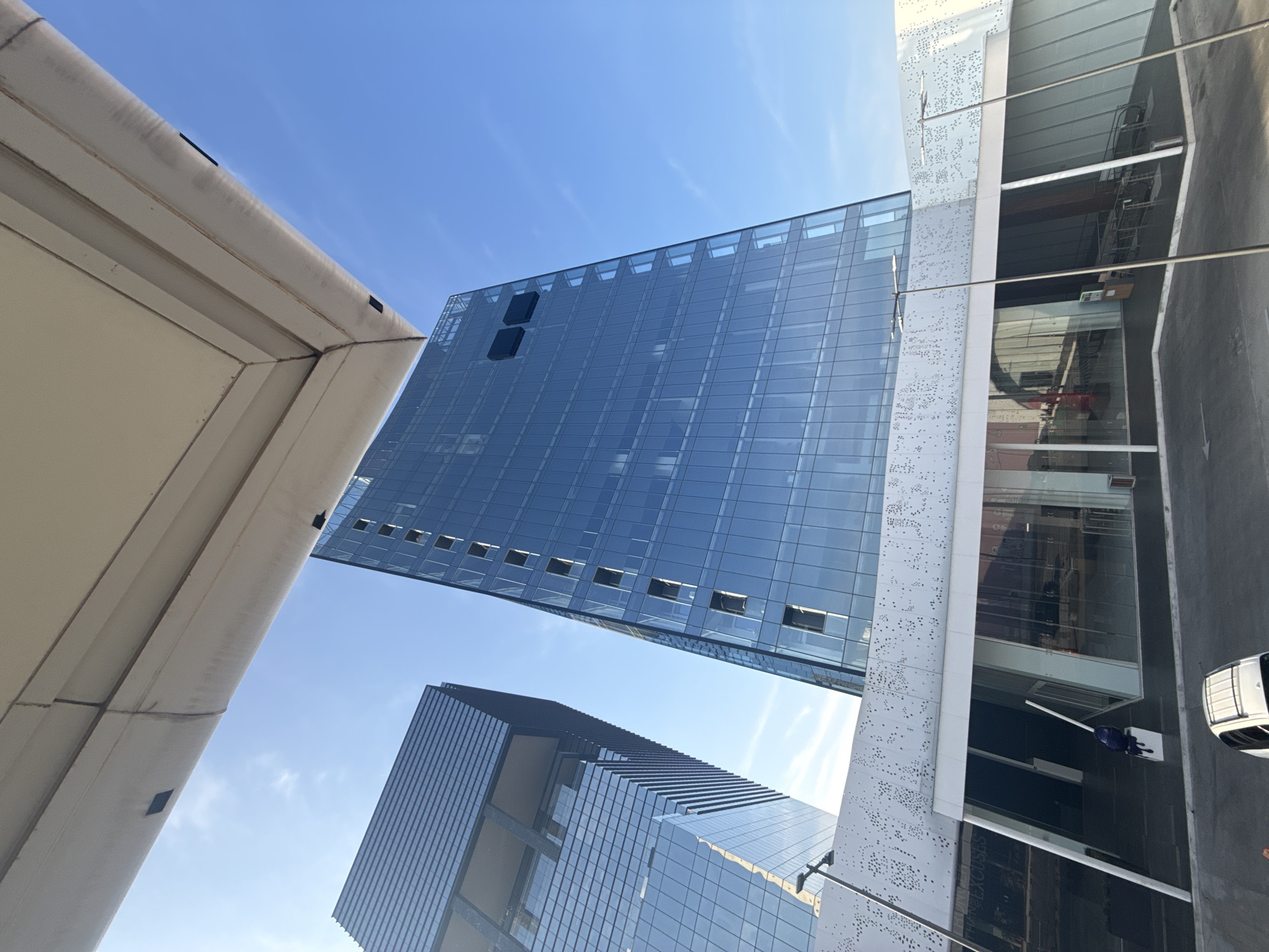The image shows a low-angle view of two tall, modern glass buildings in Puebla, Mexico. The building on the left appears to be a simple rectangular prism, while the building on the bottom-right is taller and has a more complex structure, seemingly divided into two separate towers. The sky is a clear blue with few clouds. In the bottom-right of the image, there is a white building with a perforated facade and a white car is seen parked on the street. The image is oriented so that the sky is on the right side of the image. There is also a section of white rooftop in the top-left of the image.