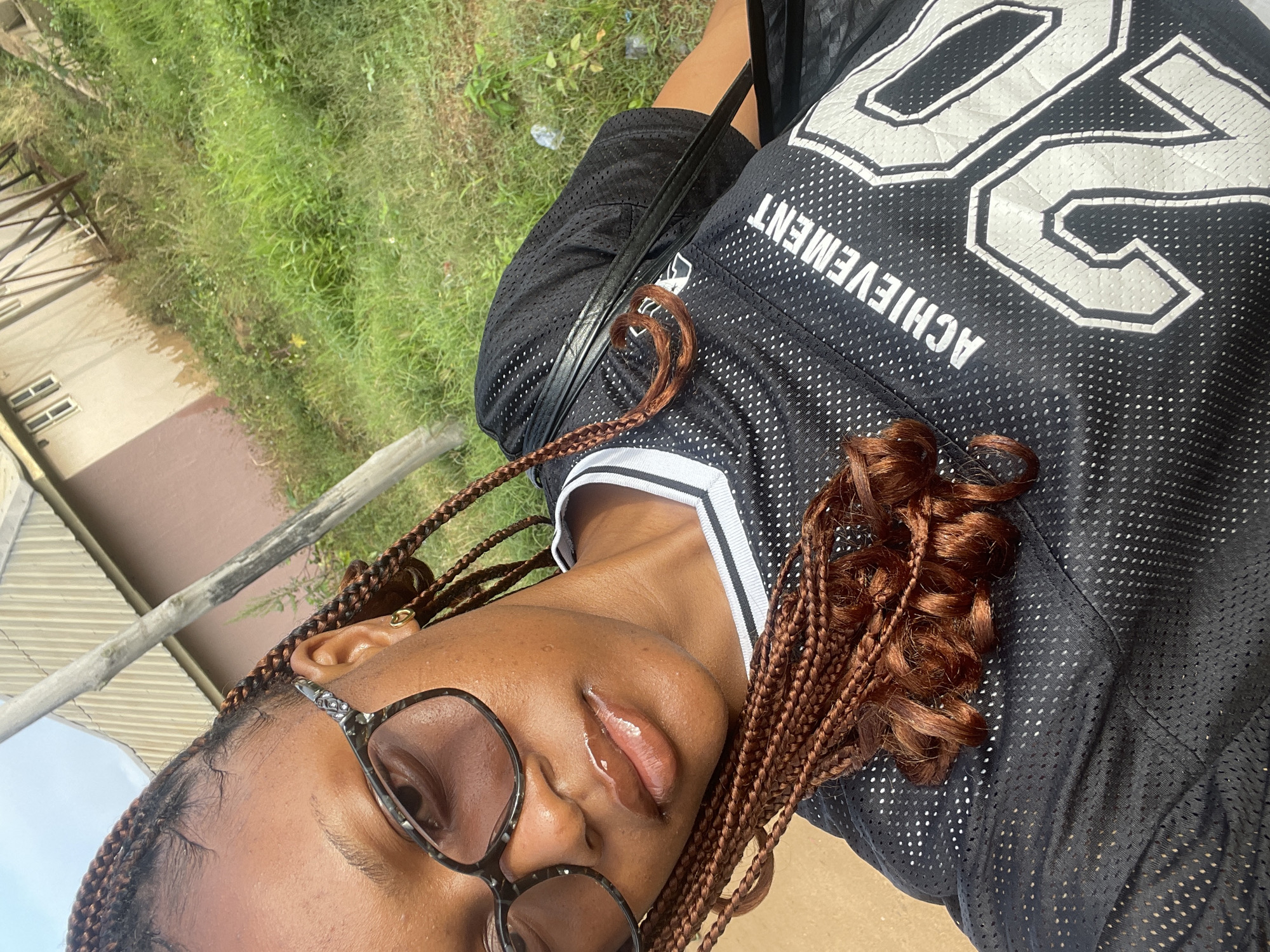 A young Black woman with braided hair, wearing glasses and a black jersey with white lettering that reads "ACHIEVEMENT 20," is pictured outdoors. She is facing the camera, with her head tilted slightly. Her lips are glossy. The background shows a building and overgrown green grass, suggesting an outdoor urban or suburban setting. The location is identified as Jos, Nigeria.