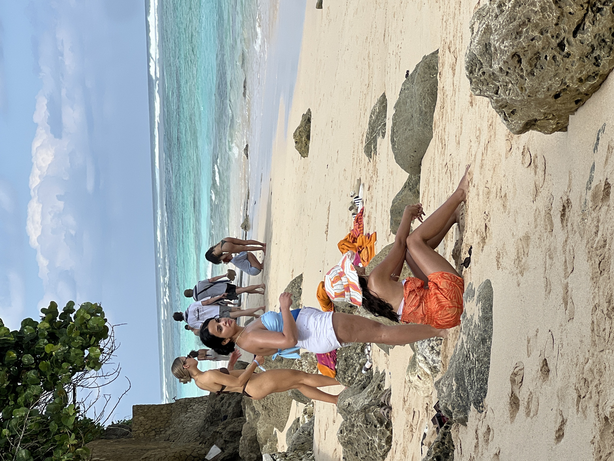 This media file captures a vibrant beach scene in Kuta Selatan, Indonesia, on a clear, sunny day. The image is oriented vertically (or rotated 90 degrees clockwise).
In the foreground, several individuals are enjoying the beach. Prominently featured are two women: one stands closer to the viewer, wearing a light blue halter top and white shorts, looking towards the camera. Another woman is reclining on the light-colored sand and rocks to the right, dressed in an orange patterned sarong or wrap over swimwear, with her dark hair splayed out. Partially visible behind the woman in blue and white is a third woman in a dark bikini, standing on a rocky outcrop.
Further down the sandy beach, towards the turquoise ocean water, several other people are standing or walking. The ocean displays clear, calm waters with gentle waves breaking near the shore. Large, textured rocks are scattered across the sandy expanse, some close to the foreground individuals, others further back on the beach. The sky above is a bright, clear blue with a few scattered white clouds. To the far left, lush green foliage from a bush or tree is visible, suggesting a natural, possibly cliff-side environment adjacent to the beach.
The overall atmosphere is one of relaxation and leisure, with people enjoying the warm weather and beautiful coastal scenery. There is no visible text on clothing, signs, or any objects within the image.