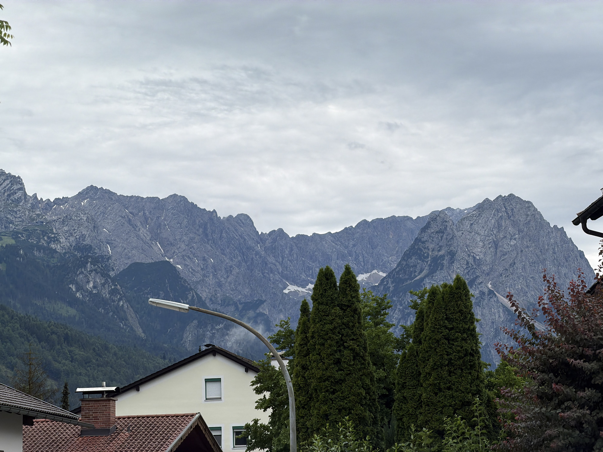 The image depicts a residential area in Garmisch-Partenkirchen, Germany, with a backdrop of imposing mountains. The foreground features the roofs of houses, one with a red tiled roof and another with a light yellow facade, a prominent brick chimney, and a modern streetlight arching over the scene. Lush green trees, including tall, conical evergreens and other foliage, partially obscure the view of the houses and the mountains.
The main subject of the image, however, is the majestic mountain range dominating the background. The mountains are rugged and rocky, with patches of snow visible on their higher slopes. The detail of the mountain range suggests a cool climate, likely late spring or early autumn. The sky is overcast with thick, gray clouds, indicating a cloudy or potentially rainy day. The lighting is diffused, casting no strong shadows, which is typical of an overcast sky.
No people are visible in the image. The overall atmosphere is serene and calm, highlighting the juxtaposition of human habitation with the grandeur of nature. The presence of houses and a streetlight suggests a developed area, likely a town or village nestled at the foot of the mountains. The combination of residential elements and the dramatic mountain landscape evokes a sense of a tranquil, alpine setting. There is no visible text in the image.
