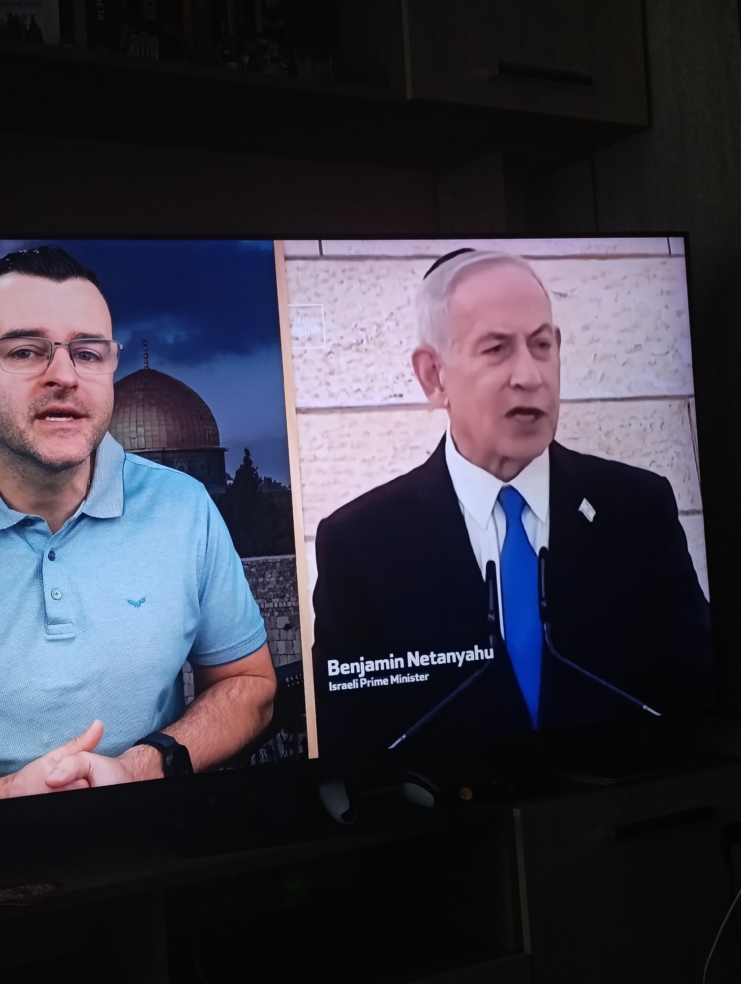 The image shows a television screen displaying a split-screen news segment. On the left is a man wearing glasses and a blue polo shirt, with the Dome of the Rock visible in the background. On the right is Benjamin Netanyahu, identified as the Israeli Prime Minister, wearing a dark suit and blue tie, speaking at a podium in front of a stone wall. The scene is shot indoors, with a dark shelf visible above the television. It is not possible to determine if this image was taken in Plovdiv, Bulgaria.