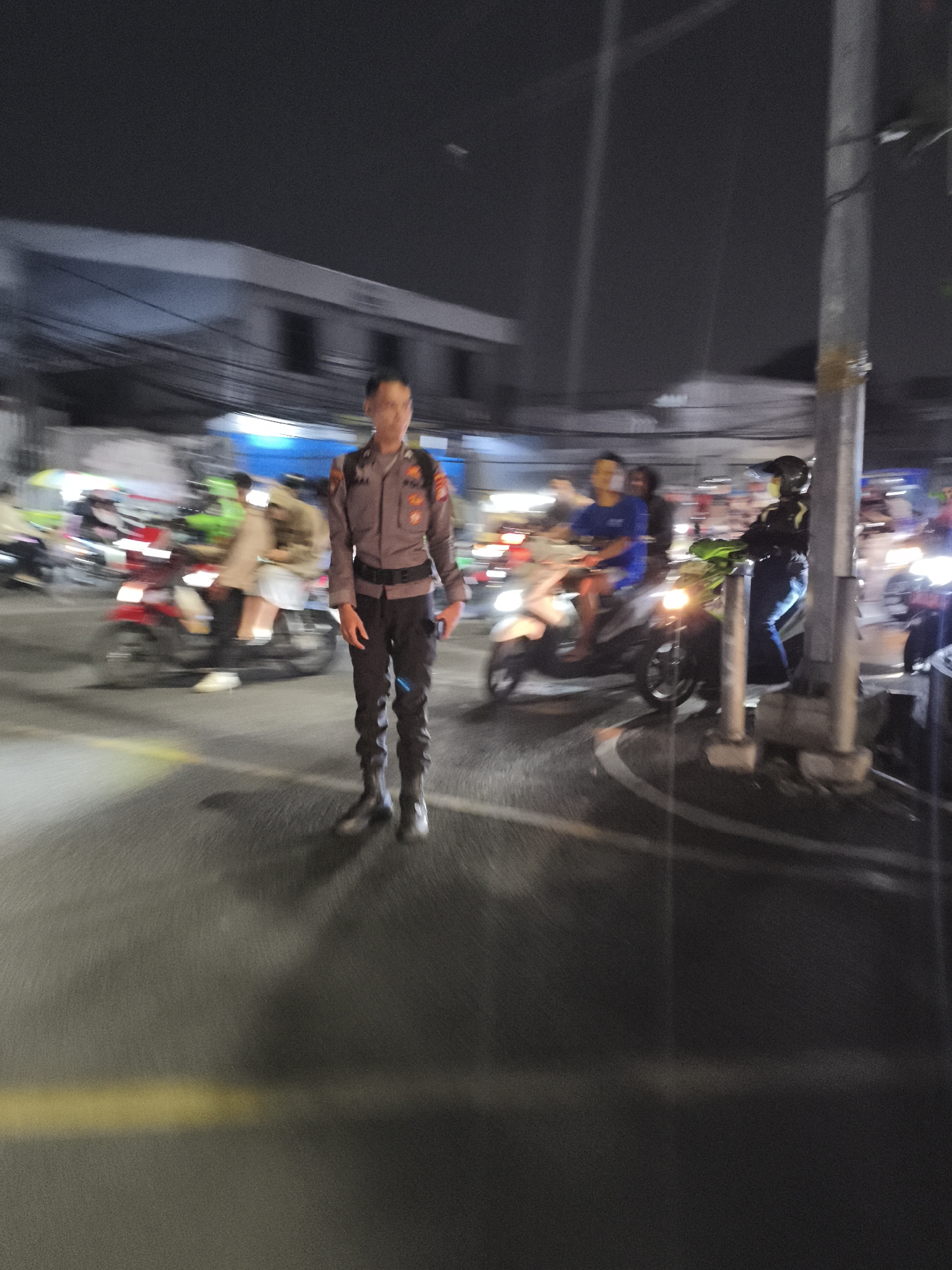 A police officer stands on a street at night in Depok, Indonesia, facing forward. The officer is wearing a dark uniform with patches on the sleeves, a belt, and boots. The background shows a street scene with several motorcycles and people, some of whom are on motorcycles. The image is slightly blurred due to movement, suggesting activity in the area. Buildings and a utility pole are also visible in the background. The lighting indicates it is nighttime.
