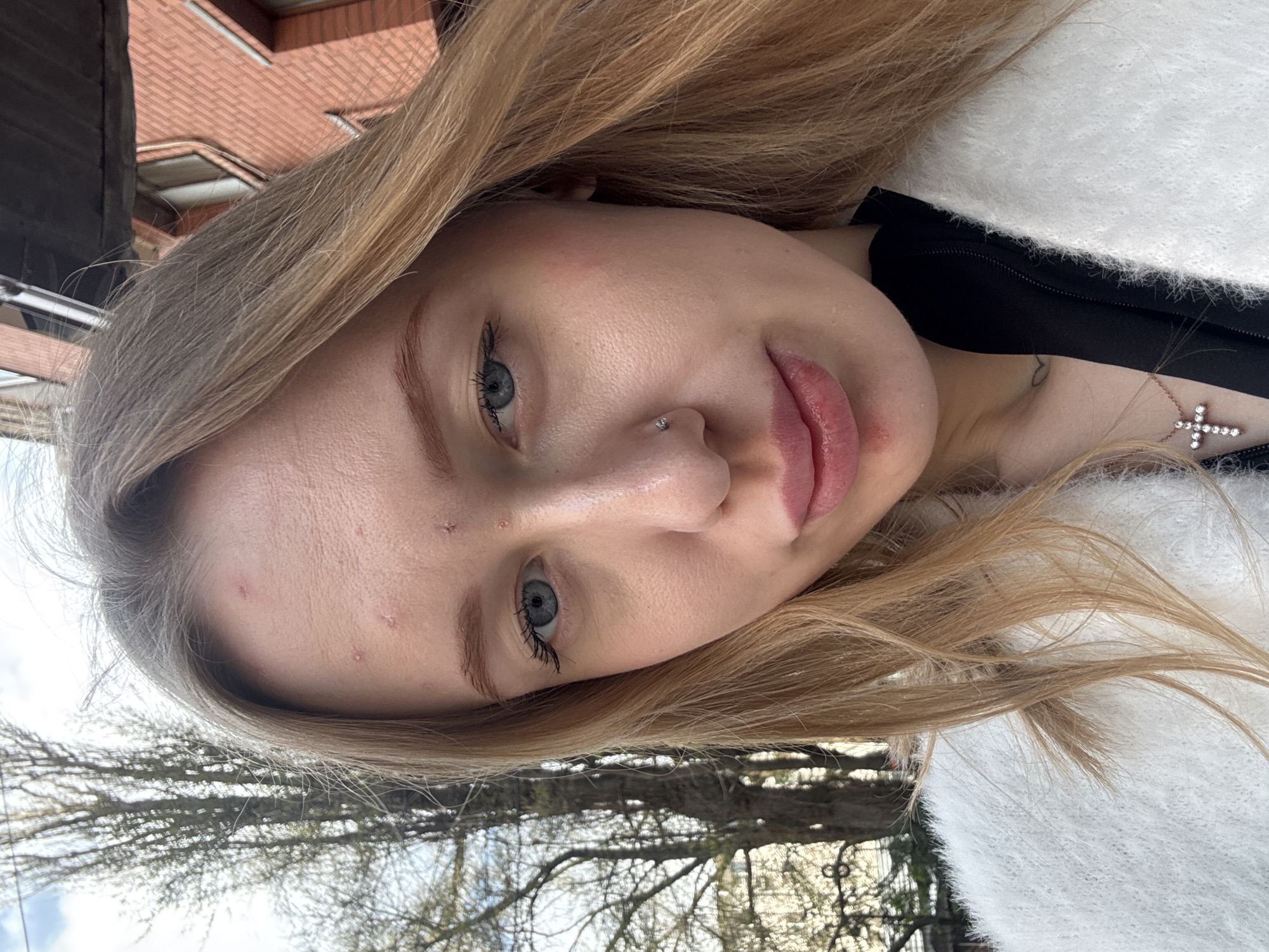 A fair-skinned woman with blonde hair is seen in a close-up shot, turned slightly to the side, with her face angled towards the camera. She has light blue eyes, a nose piercing, and visible acne scars on her forehead. She wears a white, fuzzy garment over a black top. A silver cross necklace is visible on her chest. The background features buildings with red brick and barren trees. The image was likely taken outdoors, and it is not possible to confirm if the image was taken in Rostov-on-Don, Russia.