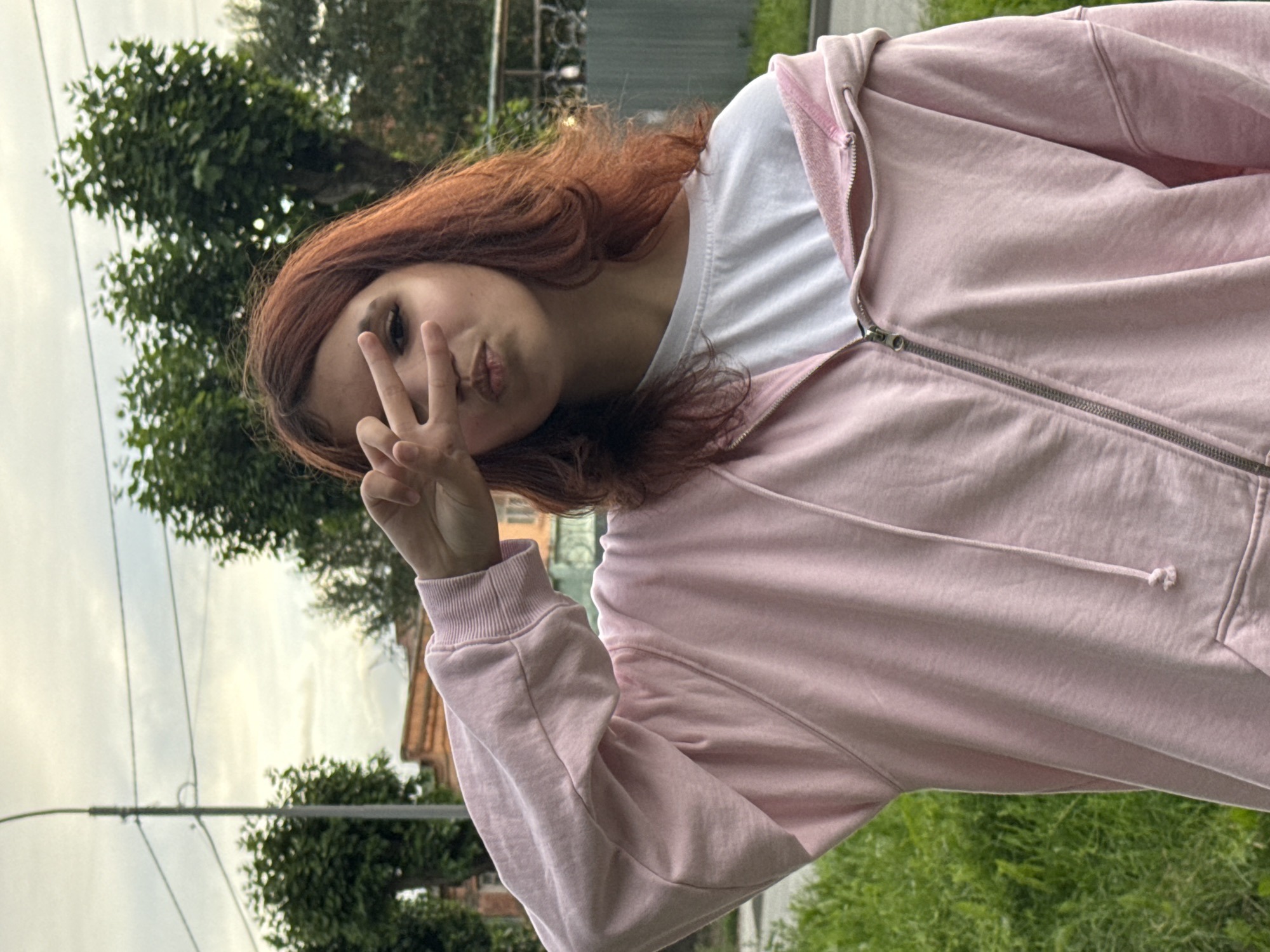 A young woman with long, reddish-brown hair is shown from the chest up, posing for a photo outdoors. She is wearing a light pink zip-up hoodie over a white t-shirt. Her face is turned towards the camera, and she is making a peace sign with her right hand, holding it up to her cheek. Her expression is playful, with her lips pursed slightly.
The background suggests a suburban or semi-rural setting. To the left, there are trees with green foliage and a glimpse of a building with a brick facade. Overhead, there are power lines against a cloudy sky, indicating overcast or twilight conditions. The overall lighting is soft. The image appears to be a selfie taken in the late afternoon or early evening. The location is Yekaterinburg, Russia, though no specific landmarks are clearly identifiable within the frame. The composition is informal, suggesting a casual moment captured for social media or personal memory.