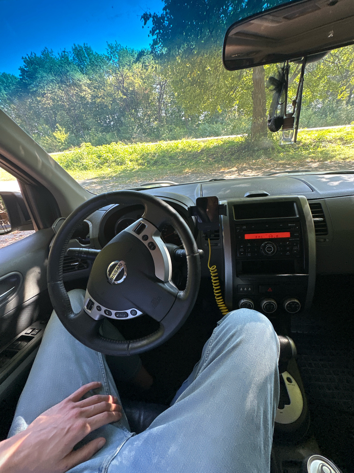 The image shows the interior of a car, likely a Nissan, with a driver's perspective. The driver's legs, clad in light blue jeans, are visible in the foreground, with one hand resting on their lap. The steering wheel, dashboard, and center console are prominent. A smartphone is mounted on the dashboard, connected to a charging cable. The car's radio display shows "FM1 90.8". Outside the car, visible through the windshield, is a sunlit, tree-lined landscape with green foliage, suggesting an outdoor setting, possibly in a rural or park-like area. The presence of a rearview mirror with what appears to be an air freshener hanging from it is also noted. The image does not contain any visual information that allows for the specific identification of the location as Shatava, Ukraine.