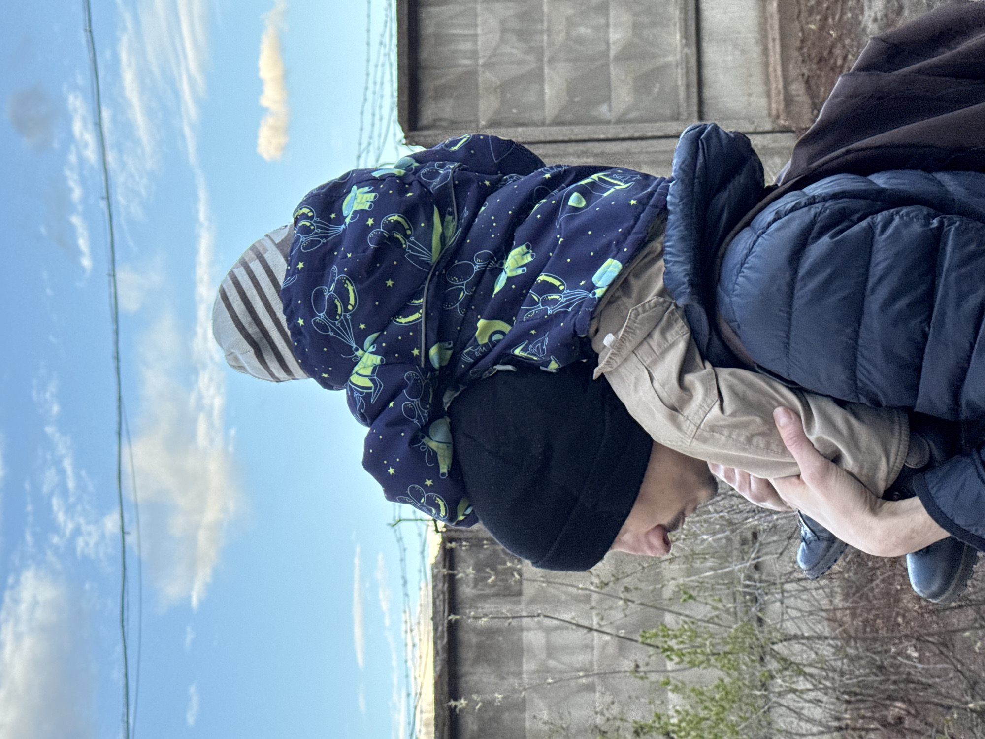 An outdoor, brightly lit image shows an adult holding a young child, appearing to carry them on their shoulders or upper back. The adult is wearing a black beanie, a dark blue padded jacket, and a light brown shirt, with their profile visible, looking downwards. Their hands support the child's legs/lower torso. The child wears a dark blue padded jacket patterned with light green/yellow balloons, stars, and stylized space-themed elements, and a striped hood or hat.
The background features a clear blue sky with scattered white clouds. A thin green wire or fence element stretches across the upper part of the sky. Below, a tall, rough concrete wall runs horizontally, topped with visible barbed wire. In the lower part of the frame, bare tree branches and some sparse green foliage suggest a transition season like early spring or late autumn. The scene takes place during daytime. No text is visible. The environment, with the concrete wall and barbed wire, suggests an industrial, perimeter, or utilitarian area within Izhevsk, Russia.