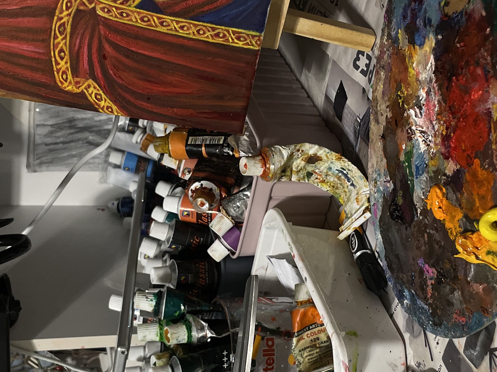 The image shows an artist's workspace, likely in a home studio. On a table, a partially finished painting on an easel depicts a figure in a red and gold robe. Beside it, various tubes of paint are arranged in containers and scattered around. A palette in the foreground is heavily laden with a variety of mixed colors, indicating active use. A white plastic tub contains more paint tubes, some of which are open. Scattered papers and tools, such as scissors, are also visible, contributing to the impression of an ongoing artistic endeavor. The lighting suggests an indoor environment, with shadows cast by overhead objects and the easel.