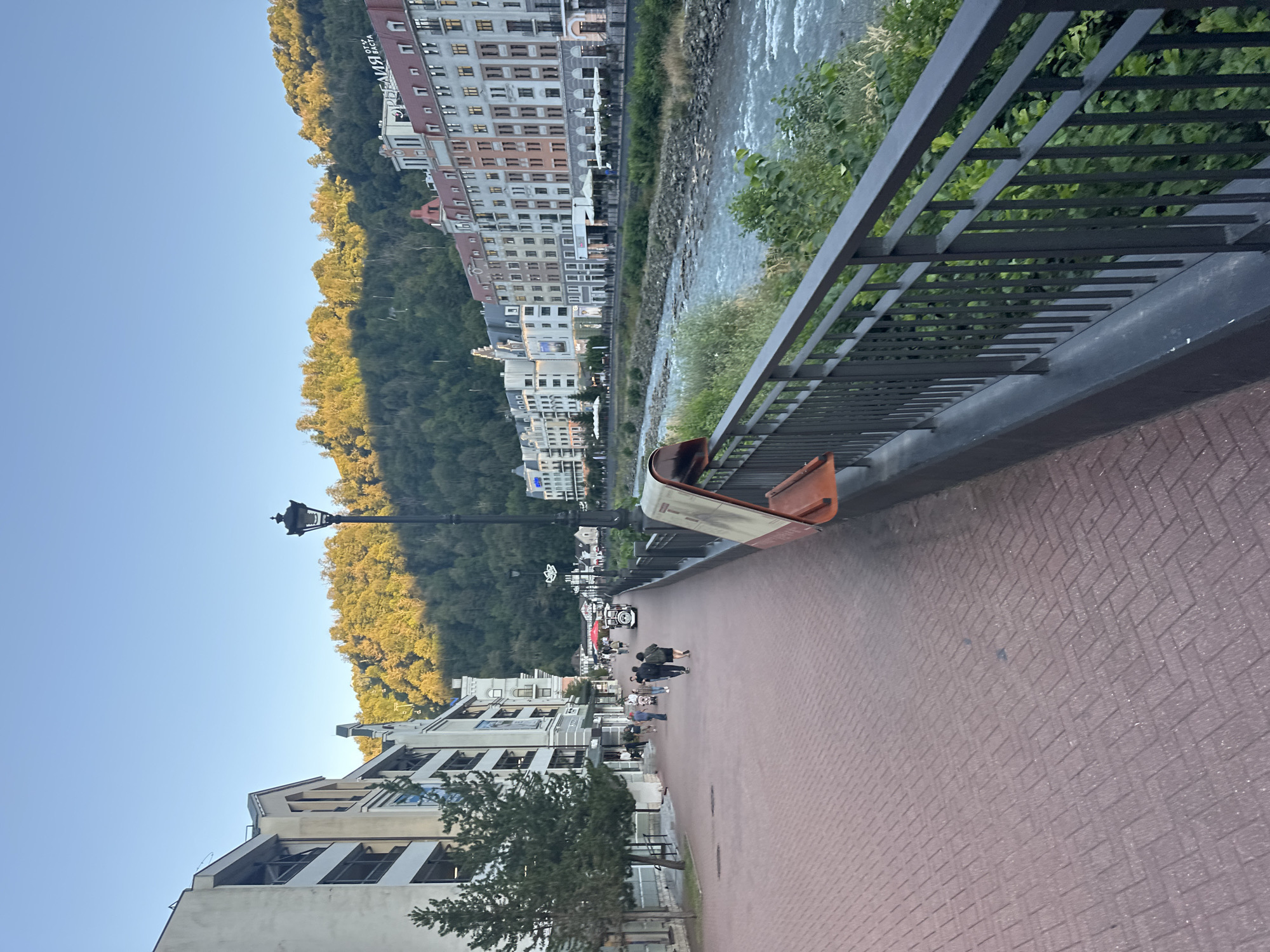 The image depicts a picturesque scene in Estosadok, Russia, along the Mzymta River. A paved walkway, constructed with red interlocking bricks in a herringbone pattern, stretches into the distance. On the left, a modern building complex with multiple stories and balconies lines the walkway, interspersed with trees. On the right, a black metal railing separates the walkway from the river. The Mzymta River flows briskly, its waters a mix of blue and grey, with rocky banks. Across the river, a row of colorful multi-story buildings, featuring pitched red roofs and uniformly spaced windows, creates a vibrant urban facade against a backdrop of lush green forested hills. The sky above is a clear, pale blue, indicating a bright, possibly late afternoon or early morning light. Several people are visible walking along the path, some individually and some in small groups. A street lamp with a traditional design stands on the walkway, and a trash receptacle with an orange surface is positioned near the railing. A white tourist train can be seen in the distance on the walkway, with people boarding or disembarking. The overall impression is one of a well-maintained, lively resort town environment.