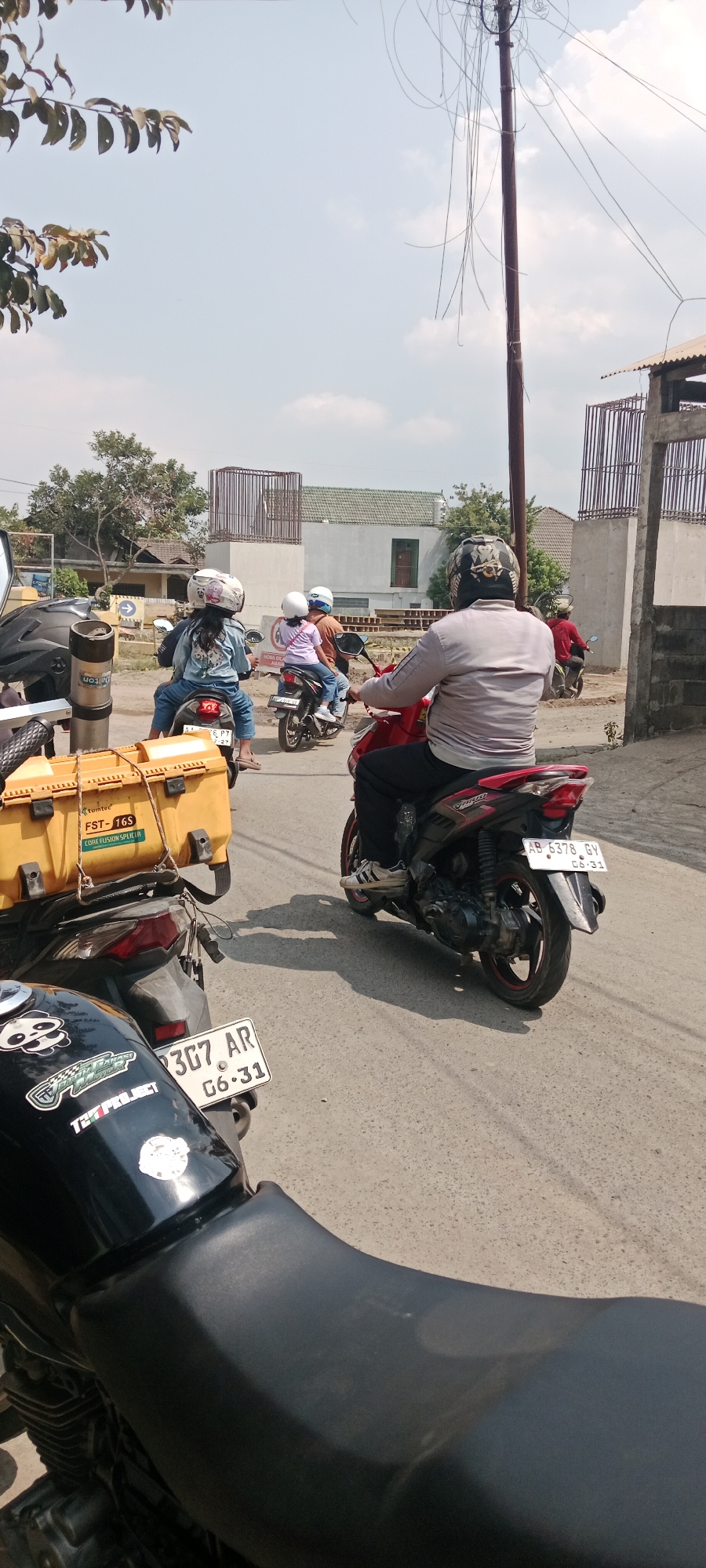 The image depicts a street scene in Purwomartani, Indonesia, on a sunny day with scattered clouds. In the foreground, a black motorcycle is partially visible, featuring a license plate that reads "3G7 AR GG-31". To its right, another motorcycle is captured from the rear, ridden by a person wearing a light gray long-sleeved shirt, black pants, and a helmet with a black and white pattern. This motorcycle has a license plate that reads "AB 5378 GY 06-31". Further ahead, two more motorcycles are visible. One is a red motorcycle carrying a person in a red shirt and a child in a purple shirt, both wearing helmets. Another motorcycle, a black one with a rider and a passenger wearing blue denim jackets and helmets, is also present. A yellow storage box is mounted on the back of one of the motorcycles in the mid-ground, labeled "FST-16S CORE FUSION SPLITTER". In the background, buildings with white walls and green roofs are visible, along with construction scaffolding. Trees and utility poles with tangled wires are also part of the environment. The road surface appears to be asphalt.