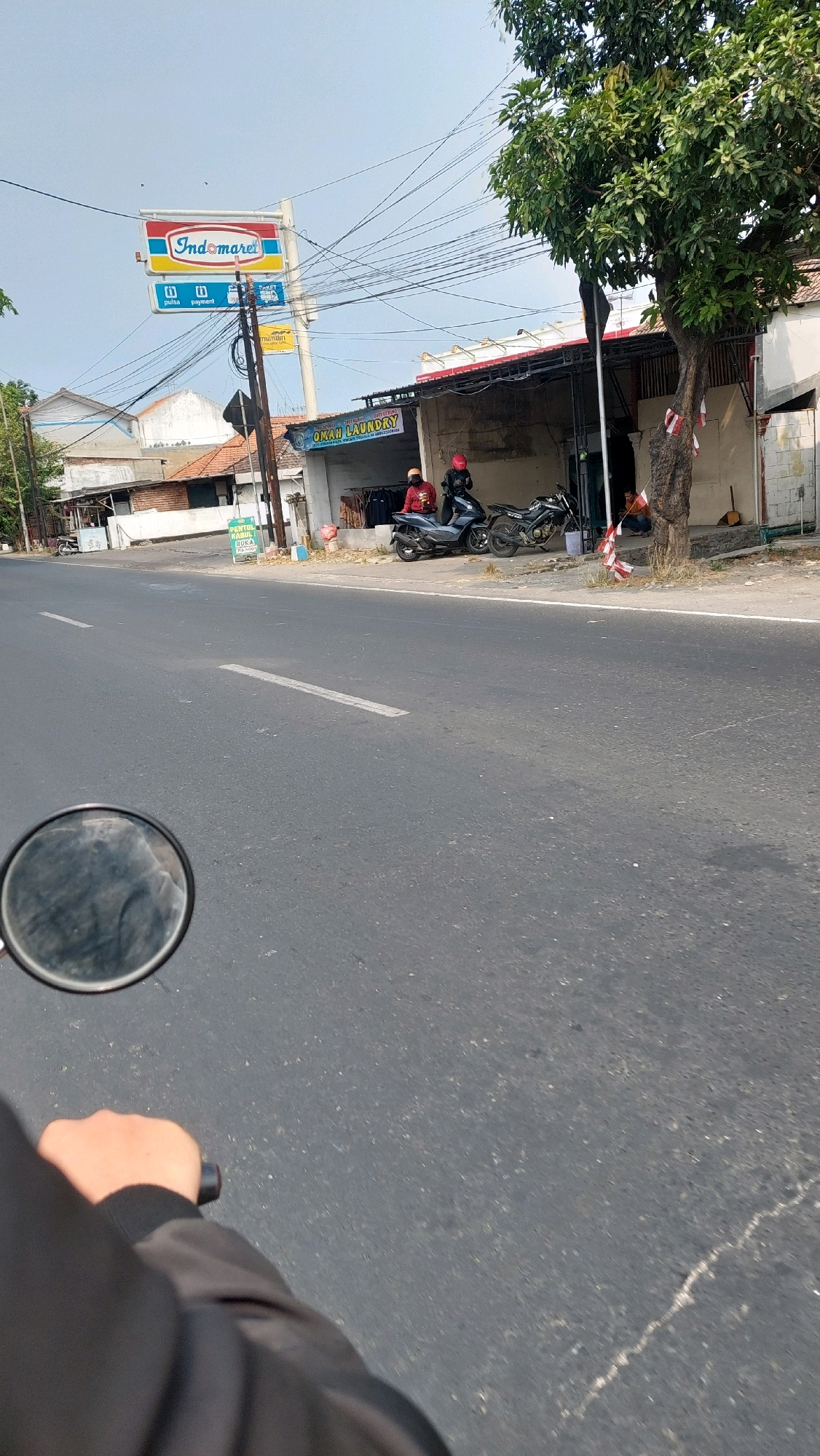 The image captures a street scene in Jimbaran Kulon, Indonesia, on a bright, clear day. In the foreground, a person's hand is visible on the handlebar of a motorcycle, with a portion of their jacket and the motorcycle's side mirror also in frame. The motorcycle is on an asphalt road with white lane markings.
To the right, a tree with green foliage stands beside a building. The building appears to be a commercial establishment, with laundry services advertised on a sign that reads "OMAH LAUNDRY". Two people are seated in front of the laundry, and two motorcycles are parked nearby. One motorcycle has a rider wearing a red helmet and another person in a red jacket is seated behind them.
Further down the street, a prominent Indomaret convenience store sign is visible, indicating its presence in the vicinity. Several utility poles with a dense network of overhead wires are also present, typical of urban landscapes. The background consists of more buildings and a clear sky. The overall impression is of a typical day in a busy Indonesian town.