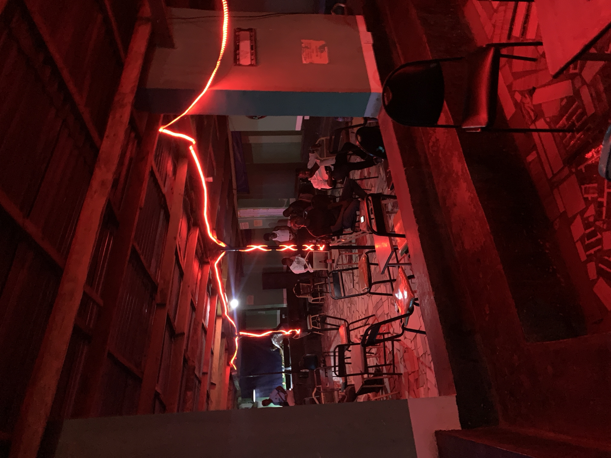 Here's a forensic description of the scene:
**Location:** Unidentified alleyway in the general vicinity of New Stadium, Nigeria.
**Environment:** A narrow outdoor alleyway, bordered by structures constructed with wood and metal sheeting on the left and what appears to be masonry on the right. The alley is dimly lit, primarily illuminated by a string of red LED lights zig-zagging overhead, contributing to a predominantly red color cast.
**Subjects/Objects:**
* **People:** Multiple individuals are visible seated at tables along the alleyway.
* **Furniture:** Tables and chairs are arranged linearly along the alleyway.
* **Lighting:** A string of red LED lights is strung overhead, acting as the primary light source.
* **Structure:** The alleyway appears to be enclosed by tall structures, one side constructed from corrugated metal and the other from masonry and tile.
* **Building details:** On the wall facing the viewer, above where people are, is a painted turquoise band. At the very top of the wall, there are two objects, probably decorative.
**Actions/Interactions:** Individuals are seated, possibly socializing or dining.
**Overall:** The scene depicts an outdoor dining or gathering area in an alleyway near New Stadium, Nigeria, marked by dim red lighting and informal seating.