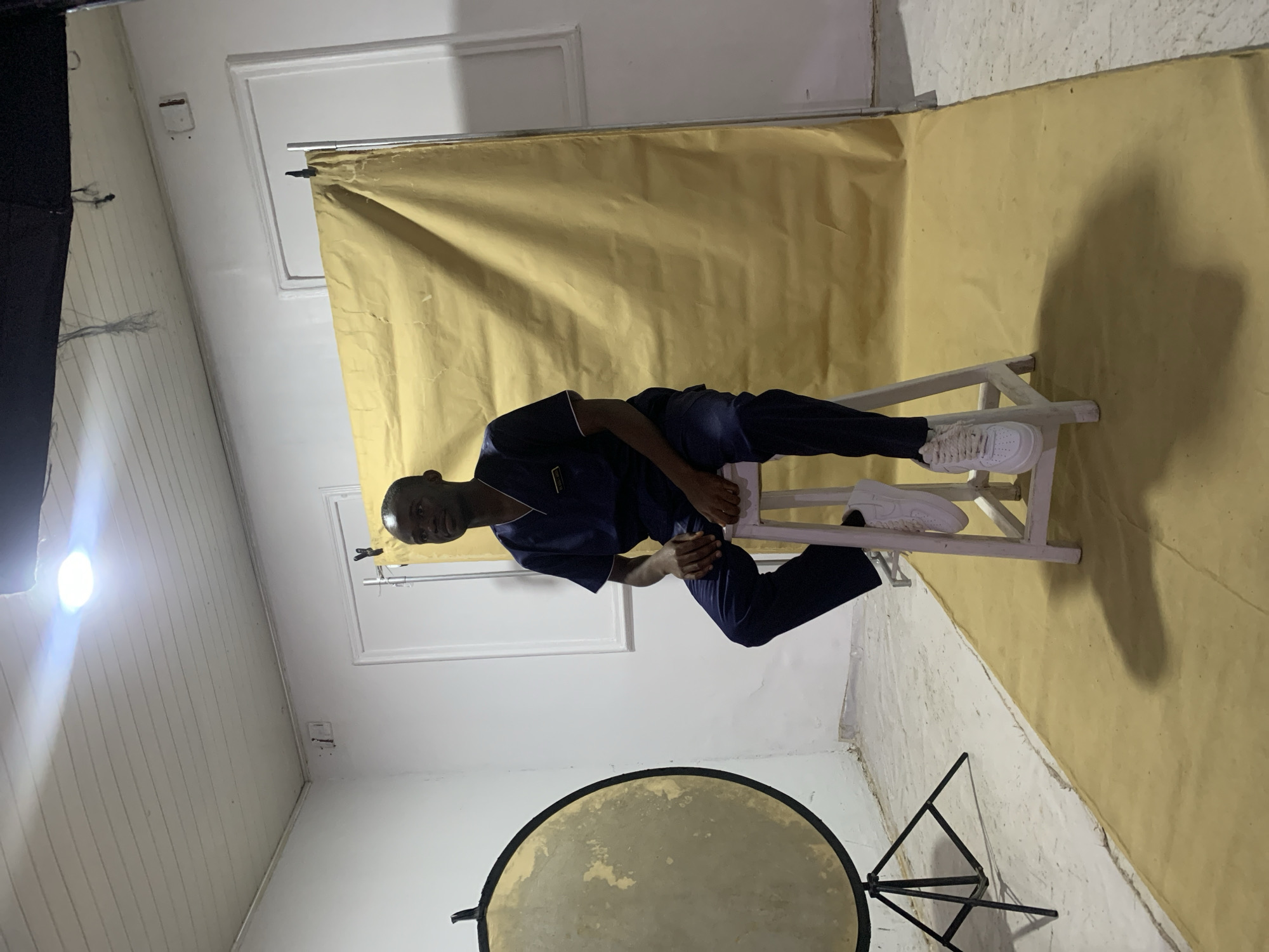 A person in a dark blue uniform and white sneakers is sitting on a white wooden chair in a room. The backdrop is a mustard-yellow sheet or backdrop. There is a reflector on a stand in the foreground, and a photography light is visible in the upper left corner. The room has white walls and what appears to be a window behind the person. The location is indicated by the style of dress, lighting equipment, and background that suggest the image was captured within a photography studio, most likely situated in a place that reflects urban life. However, I don't have access to information about the interior of Jos, Nigeria. Therefore, I'm unable to pinpoint the exact location.