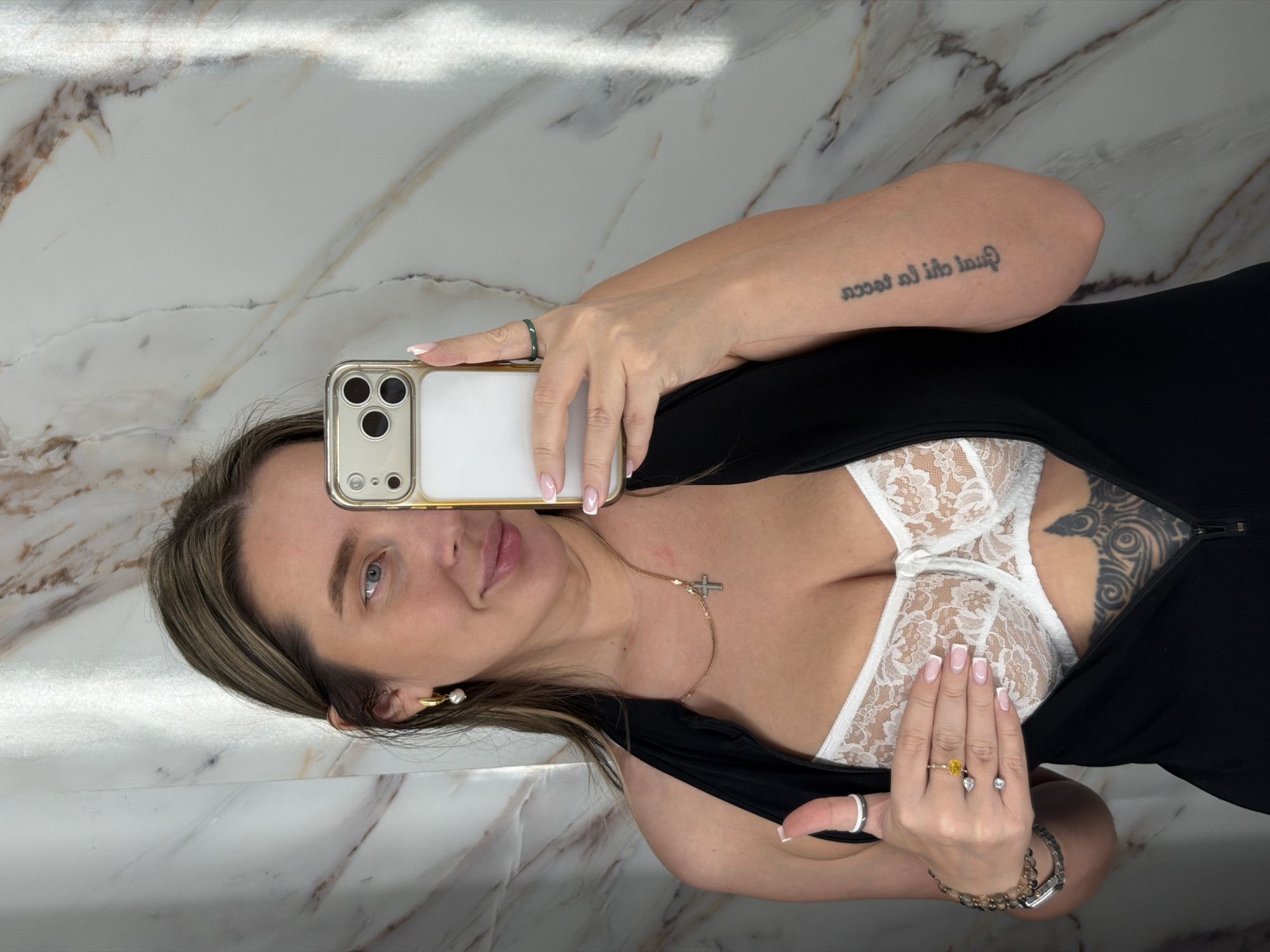 A woman is posing for a selfie in front of a marble-patterned wall. She is holding a smartphone with a gold-colored case in her left hand, partially obscuring her face. She has blonde hair with highlights, fair skin, and light-colored eyes. She is wearing a white lace bra and a black top or dress. She has a tattoo on her left arm and another on her chest near her right breast. She is wearing jewelry, including rings, a gold necklace with a cross pendant, and earrings. She also has a bracelet on her right wrist. The location is not identifiable as Moscow, Russia, based on the visual information provided.