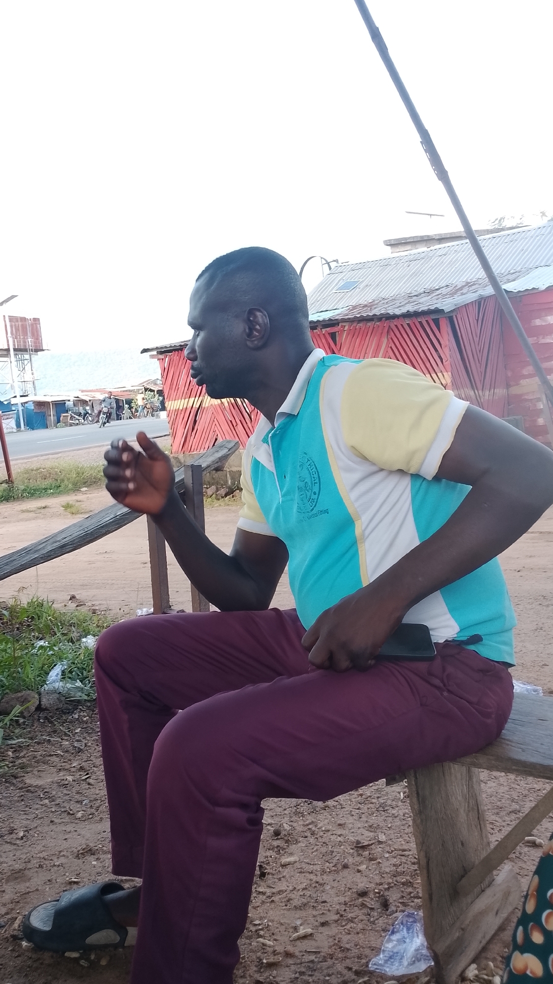 An African man in his 30s, with dark skin and a shaved head, is seated on a wooden bench. He is wearing a light blue and yellow polo shirt with a logo on the left side of his chest and burgundy pants. His left arm is bent at the elbow, with his hand open as if in conversation or gesturing. His right hand rests on his lap, holding a dark-colored smartphone. The man is looking to his left, away from the camera.
The background consists of a red building with a corrugated metal roof, likely a shop or small structure. To the left, a road is visible with a few motorcycles and people. The ground is covered in dirt and scattered debris. The overall setting appears to be an outdoor urban or semi-urban environment, consistent with a location in Adankolo, Nigeria. The sky is overcast.