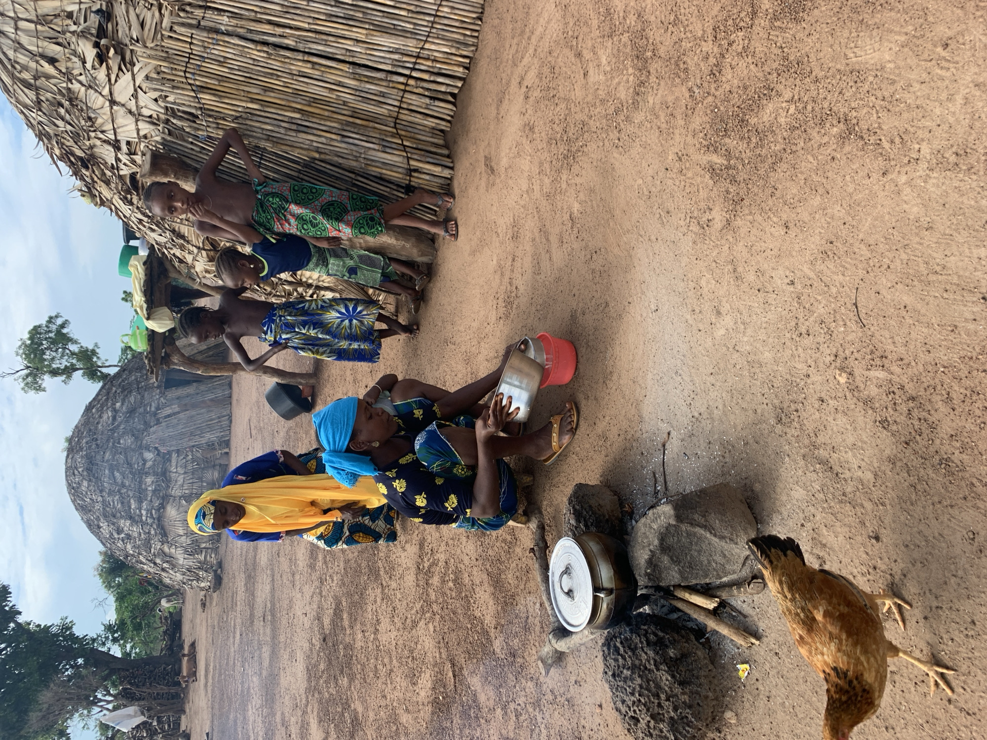 In Nandu, Nigeria, a group of individuals are gathered in a village setting. A woman wearing a blue headscarf and a patterned top is crouched down, attending to a pot cooking over an open fire made of sticks and stones. She holds a metal bowl in her hands. Next to her, another woman in a yellow headscarf and blue and yellow patterned attire stands. Three children, two girls and a boy, are also present. The children are barefoot and wear colorful patterned skirts. The boy stands slightly apart, while the girls are closer to the woman with the pot. Behind them, a conical hut constructed from thatched grass and bamboo stands, its entrance visible. Another similar structure is partially visible in the background to the left, along with trees and vegetation. A chicken is walking on the sandy ground in the foreground. The scene depicts daily life and activities within a rural Nigerian community.