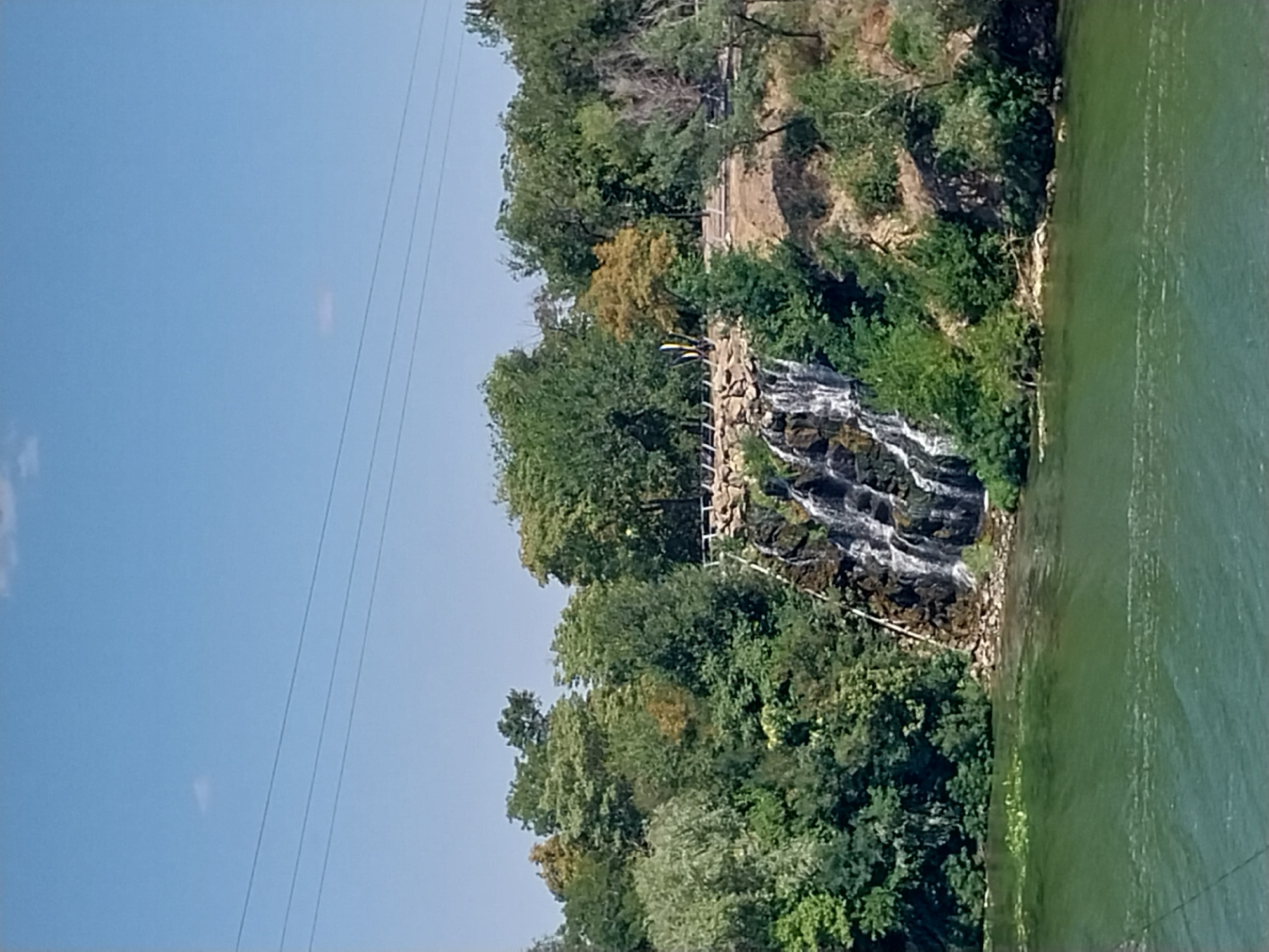 The image shows a man-made waterfall cascading down a rocky embankment into a body of green water. The waterfall is surrounded by lush green trees and vegetation. A pathway runs along the top of the embankment, with a white railing. In the distance, there are power lines stretching across a clear blue sky. The scene appears to be a park or recreational area. This is located in the city of Dnipro, Ukraine.