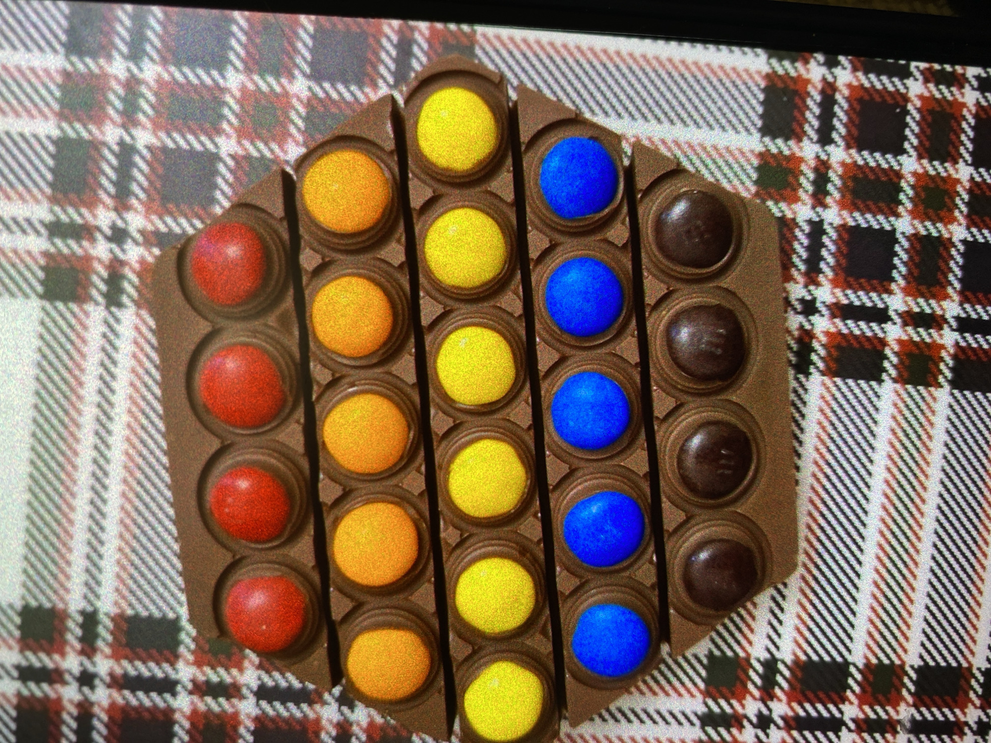The image displays a hexagonal-shaped chocolate bar, designed to resemble a pop-it fidget toy. It is divided into multiple individual compartments, each holding a colorful, candy-coated chocolate or a plain chocolate button. The candies are arranged in distinct vertical columns by color: red on the far left, followed by orange, then bright yellow, and finally blue. The rightmost column contains plain brown chocolate buttons. The chocolate bar itself appears to be milk chocolate.
The chocolate is placed on a surface that exhibits a plaid or tartan pattern. This pattern, featuring interwoven lines of white, black, and red, with some subtle grey/dark tones, appears to be a digital image displayed on a screen, indicated by visible pixelation and moiré effects, particularly evident in the cropped close-ups.
The scene is a static arrangement, suggesting a product photograph or a still-life display. There is no activity occurring, and no individuals are present. The chocolate and candies are in perfect condition, showing no signs of being consumed or tampered with. No time of day, specific weather conditions, or emotional states can be inferred. There is no visible text within the image. While the contextual information points to Saint Petersburg, Russia, the visual content of the image does not provide any specific cues to this location.