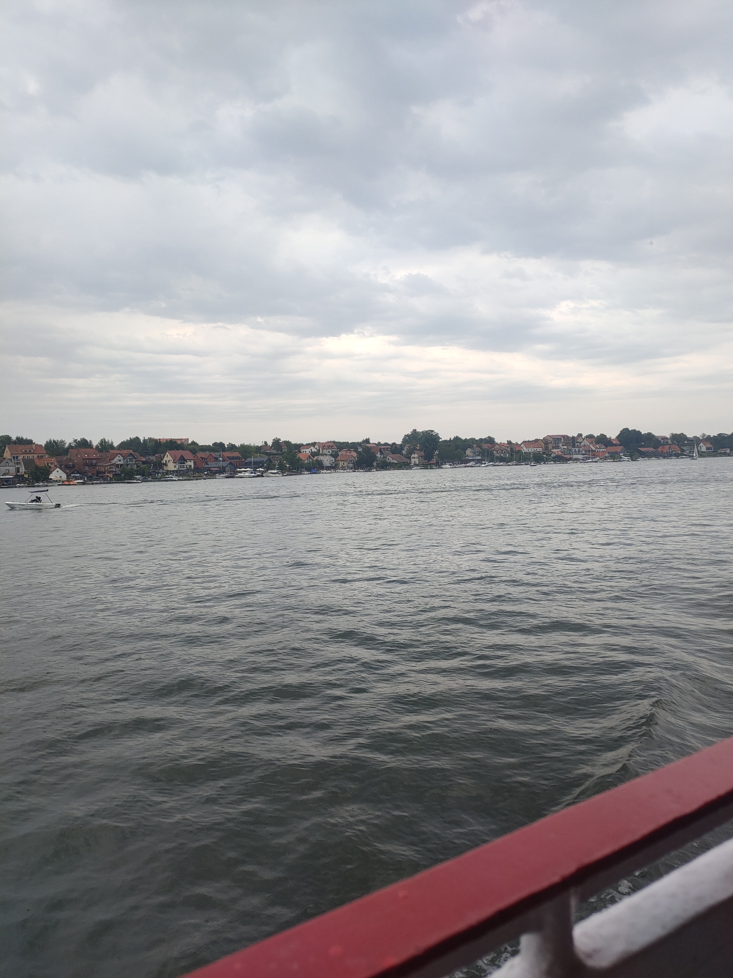 A motorboat is moving through the gray water of a lake towards a shoreline with numerous houses and trees. The sky is overcast with clouds. The foreground of the image shows the railing of a boat, painted red and white.