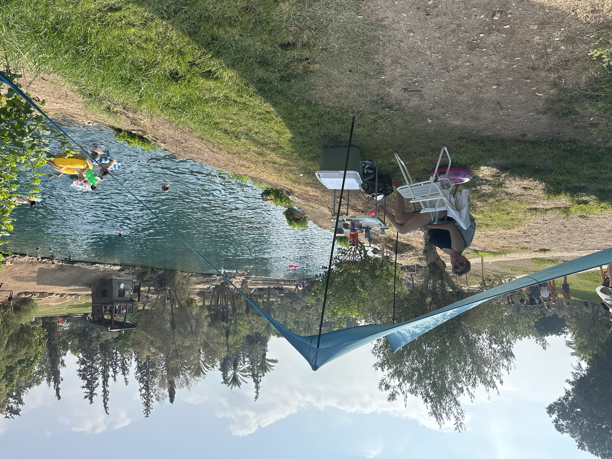 The image depicts a scene at the Sachne National Park, located in the Emek HaMaayanot Regional Council, Israel. The park features a clear, turquoise-blue body of water, likely a natural pool or springs, surrounded by lush green vegetation and trees, including palm trees and olive trees. The sky is partly cloudy, indicating a bright, sunny day.
In the foreground, a woman is seated on a white folding chair under a blue canopy tent. She is wearing a black tank top and shorts, with sunglasses on her face. Next to her, a small table holds various items, including drinks and snacks. A green and white cooler is placed on the grass nearby.
In the water, several people are enjoying themselves. Some are swimming, while others are relaxing on inflatable tubes, including a large yellow pineapple-shaped tube and a striped blue and white tube. A wooden lifeguard tower stands on the bank of the water, to the right of the frame. In the background, a group of people, including children, are visible walking along a path or standing near the water's edge. The overall atmosphere suggests a recreational outing, possibly a picnic or a day trip to swim and relax.