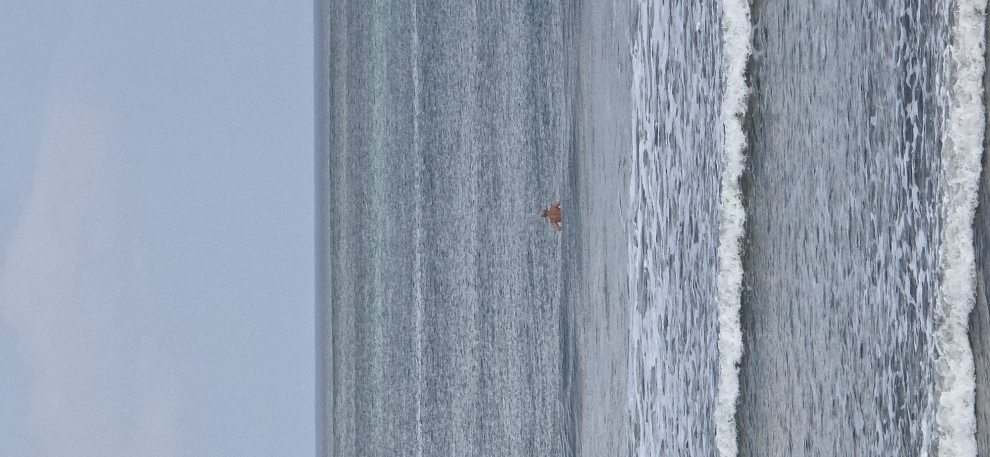A solitary man stands waist-deep in the ocean with his back to the camera. He has fair skin and appears to be bald or have very short hair. He is wearing orange shorts. The water is a muted gray-blue, and gentle waves are breaking in the foreground. The horizon is clearly defined, separating the ocean from a pale, overcast sky. The scene appears to be a typical day at the beach in Kuta, Indonesia, characterized by its expansive shoreline and moderate ocean conditions.