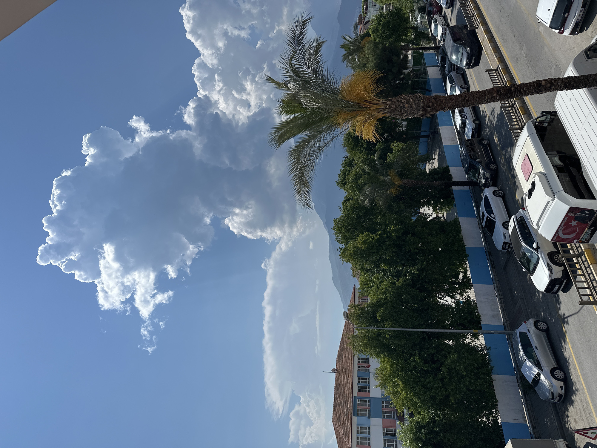 The image shows a daytime scene in Fethiye, Turkey. The view appears to be from a high vantage point, looking down onto a street with parked vehicles.
Sky: A large, puffy cumulus cloud dominates the left side of the image. The sky is blue.
Landscape: Mountains are visible in the distance, appearing hazy and light in color. There are trees and green foliage visible along the street. A palm tree stands out near the center of the image.
Architecture: A building with a red tile roof is partially visible on the left side.
Street Scene: A line of parked cars and vans extends along the street. A blue and white painted barrier is adjacent to the street.
Objects: A ladder is leaning against a white van. The Turkish flag is visible on a van.