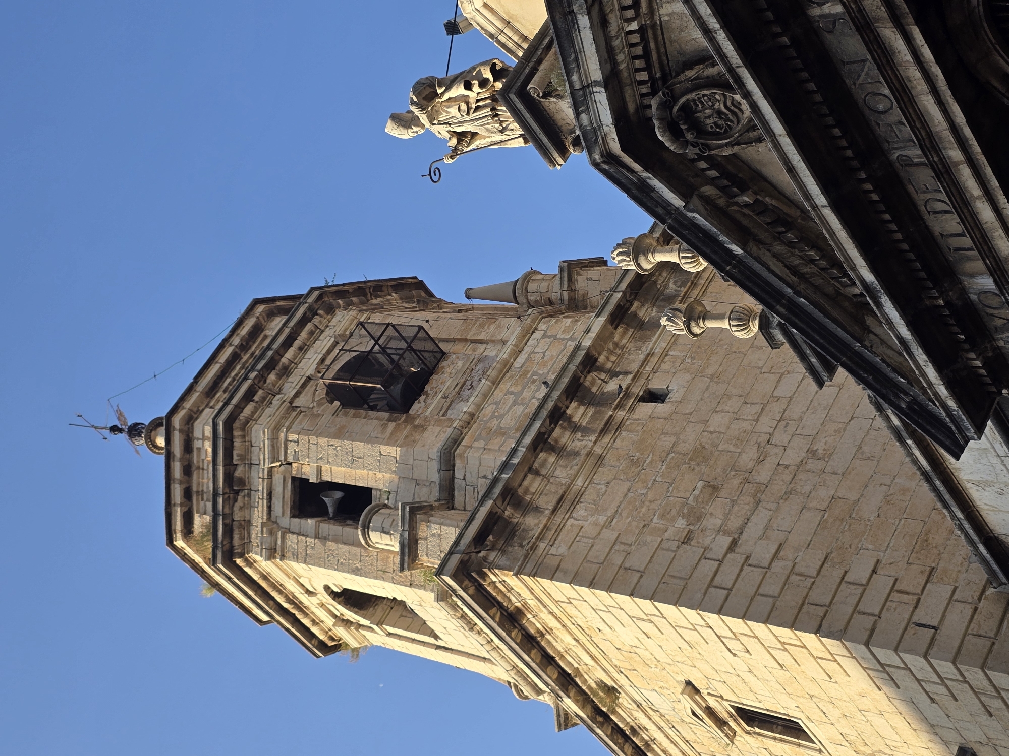 This is a low-angle shot of the bell tower of the Jaén Cathedral, located in Jaén, Spain. The stone tower, constructed from roughly cut blocks, rises against a clear blue sky. Visible features include arched openings, likely for bells, some of which appear empty or have metal grates. A weather vane is perched atop the tower. To the right of the tower, the upper facade of a building is visible, featuring an ornate cornice and a statue of a robed figure holding a staff. The statue is positioned on a pedestal above a carved crest. The overall impression is of historical architecture under bright daylight.