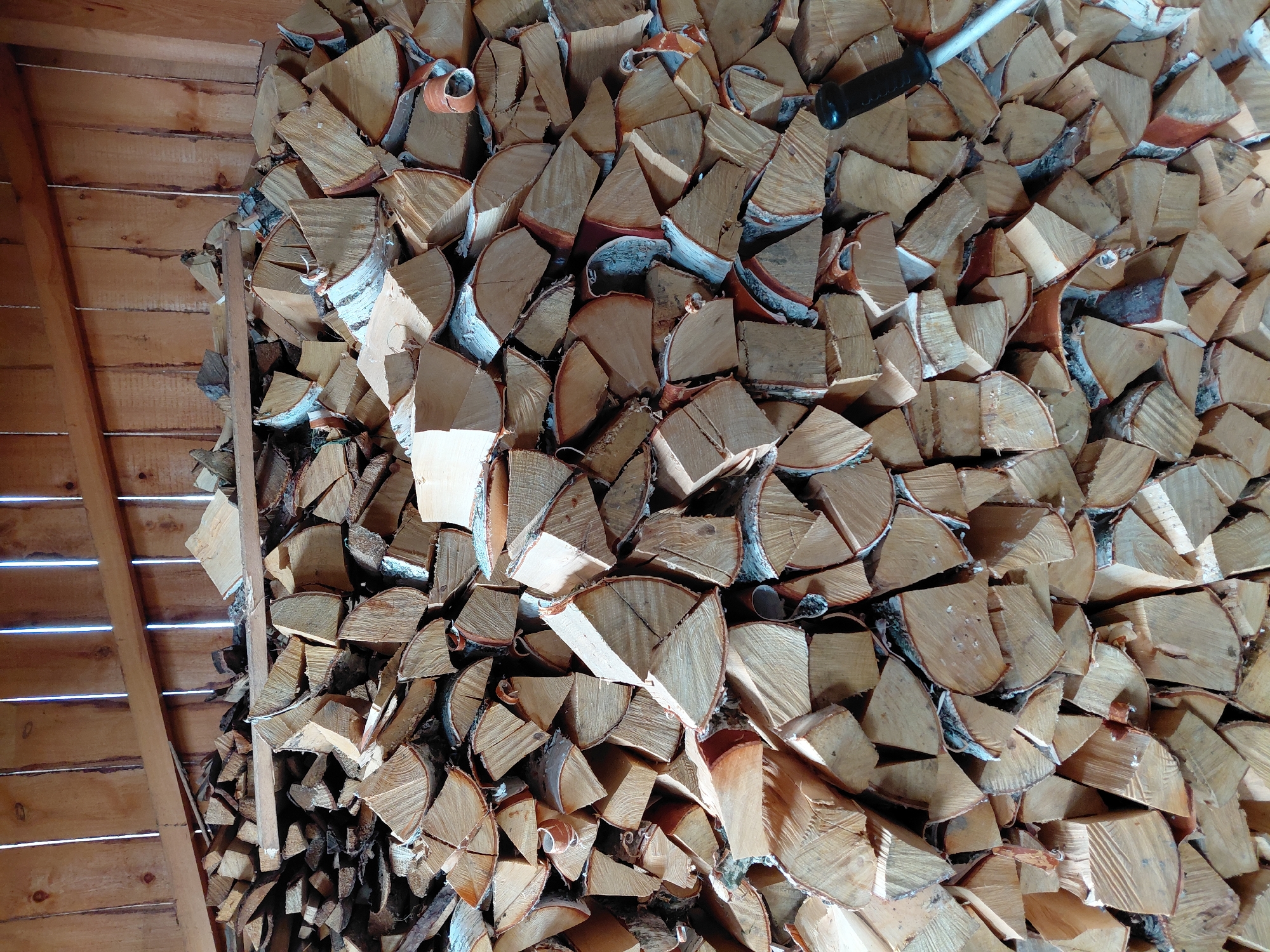 A stack of split firewood is stored inside a wooden structure. The firewood is predominantly birch, with visible white bark and light-colored wood. The logs are cut into various lengths and shapes, with some exhibiting a more uniform, split appearance and others showing rougher edges. The wood is piled high against a wooden wall, which is constructed of vertical planks with visible grain and knots. Light streams in from the top of the structure, creating highlights and shadows on the wood. A black handle of a tool, possibly a shovel or rake, is visible on the right side of the image, extending partially into the pile of firewood. The overall scene suggests a practical, rustic setting for firewood storage. The context of Malinovka, Russia, implies a cold climate where such firewood would be essential for heating.