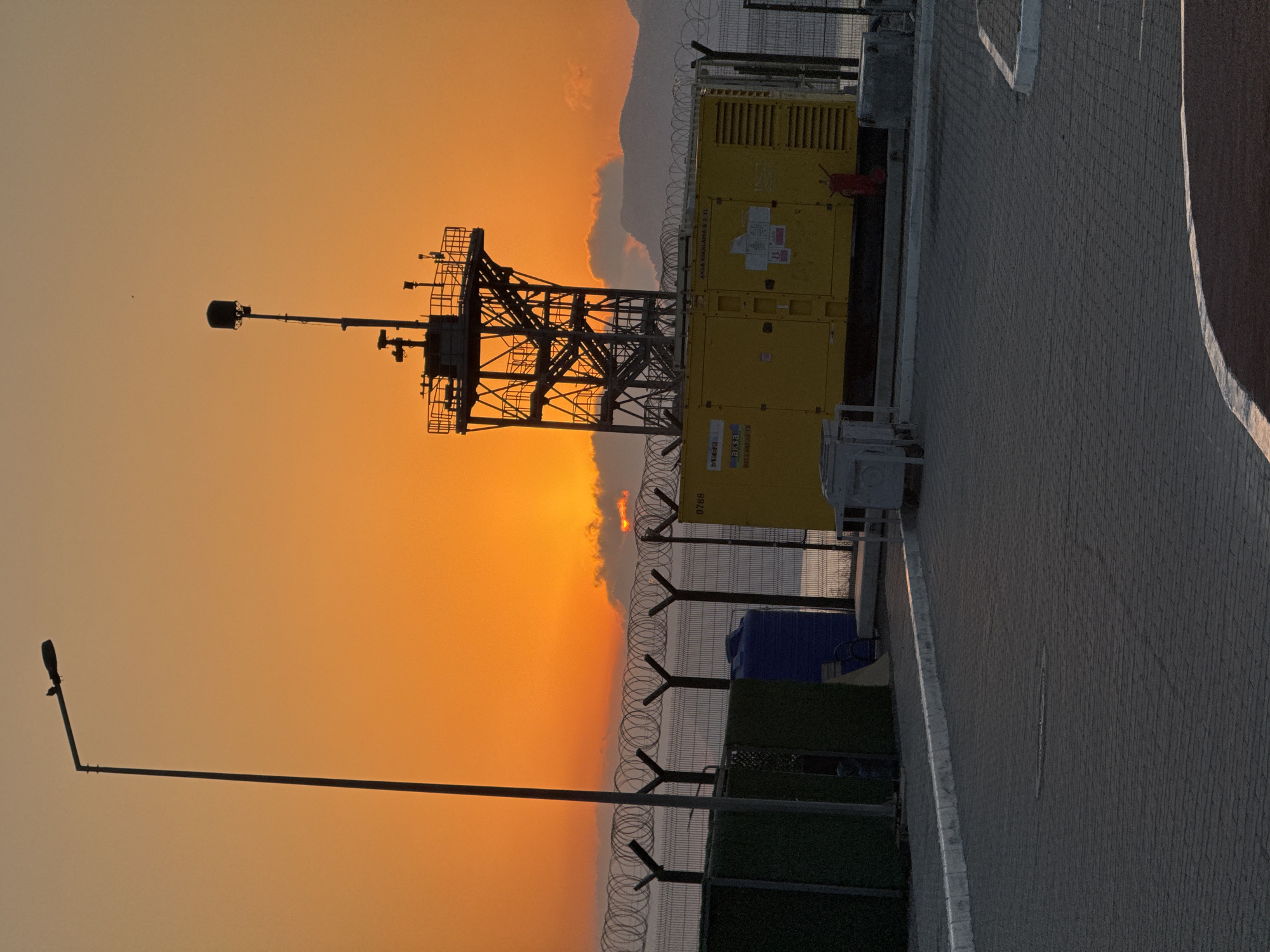A wide-angle, eye-level outdoor shot captures a sunset over a coastal area in Gülnar, Turkey. The sky is a vibrant gradient of orange and yellow, with a few wispy clouds. In the foreground, a paved area transitions to a gravelly section. To the left, a dark green fence encloses a small structure. A tall, thin lamppost stands prominently in the left midground, silhouetted against the sky, with a bird perched on its arm.
In the midground, a chain-link fence topped with razor wire separates the foreground from the background. Behind the fence, a large yellow generator sits. To the left of the generator is a blue water tank. Further back, a dark, mountainous landscape is visible, partially obscured by clouds. A tall, metal tower structure with stairs and railings rises behind the generator. The scene is devoid of any people.
