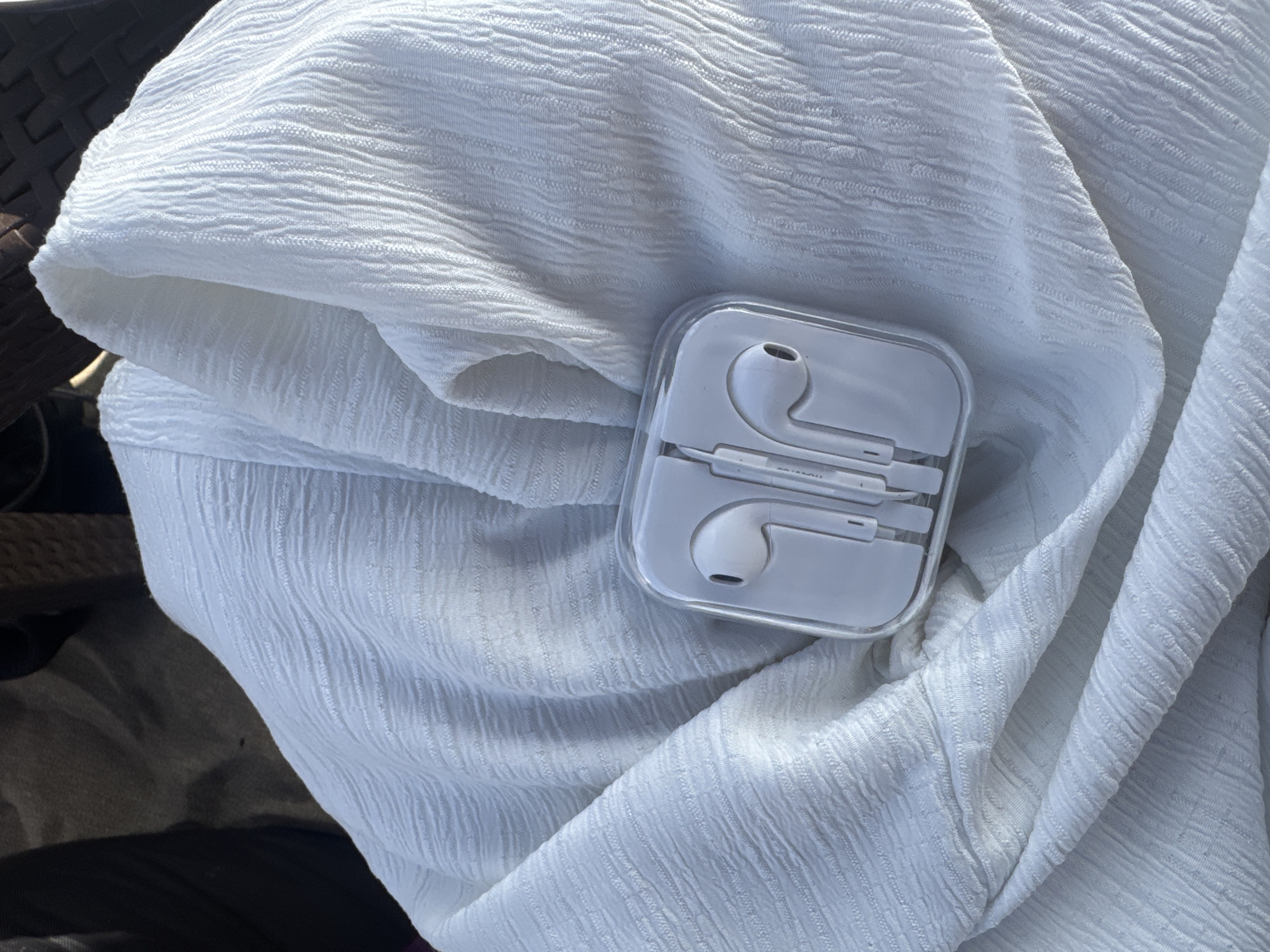 A pair of white earphones in a clear plastic case are placed on a white textured fabric. The fabric is draped and folded, creating shadows and highlights. In the background, out of focus, are dark brown and black objects, possibly furniture or clothing. The scene appears to be outdoors due to the lighting and the sandy texture of what appears to be the ground beneath the fabric. The image doesn't contain any specific identifiable landmarks or elements that would indicate it is in Kuta, Indonesia.