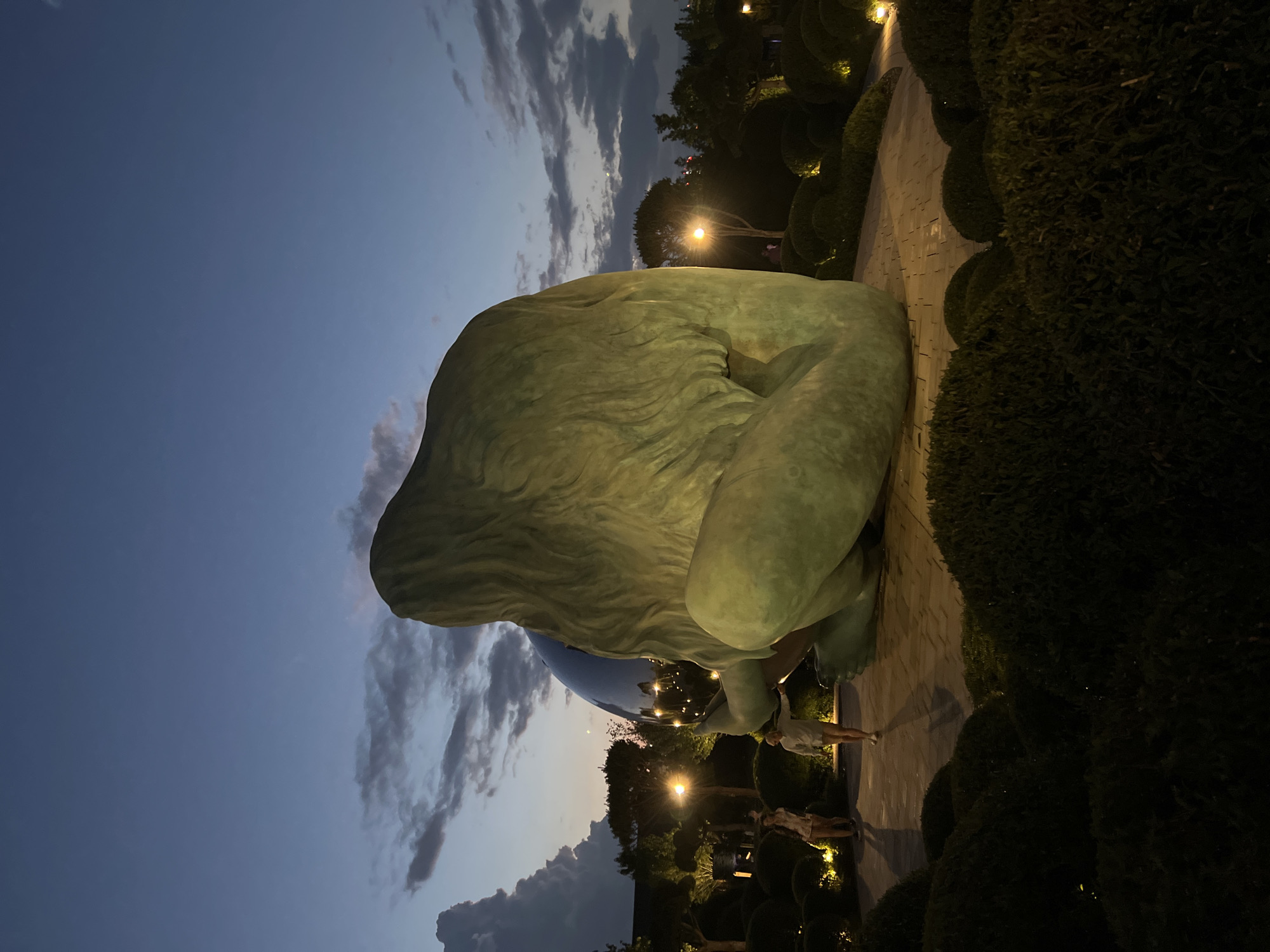 This image shows a large, green, abstract sculpture resembling a hunched figure outdoors at dusk. The sculpture is positioned on a paved walkway, flanked by neatly trimmed, dark green bushes that are illuminated by warm outdoor lights. The sky above is a deep blue with scattered clouds, indicating twilight.
In the background, there are more trees and sculpted bushes, along with streetlights casting pools of light on the path. Two people are visible near the sculpture, one standing and the other walking. They appear small in comparison to the massive artwork, suggesting its scale. The lighting creates a somewhat dramatic and serene atmosphere.
The location is identified as Krasnodar, Russia. The overall impression is of an art installation in a park or public space, enjoyed during the transition from day to night. The sculpture's form is organic and rounded, and its surface has a textured, weathered appearance. The reflective sphere beneath the sculpture's chin adds a contrasting element of polished modernity to the otherwise earthy tone of the main piece.