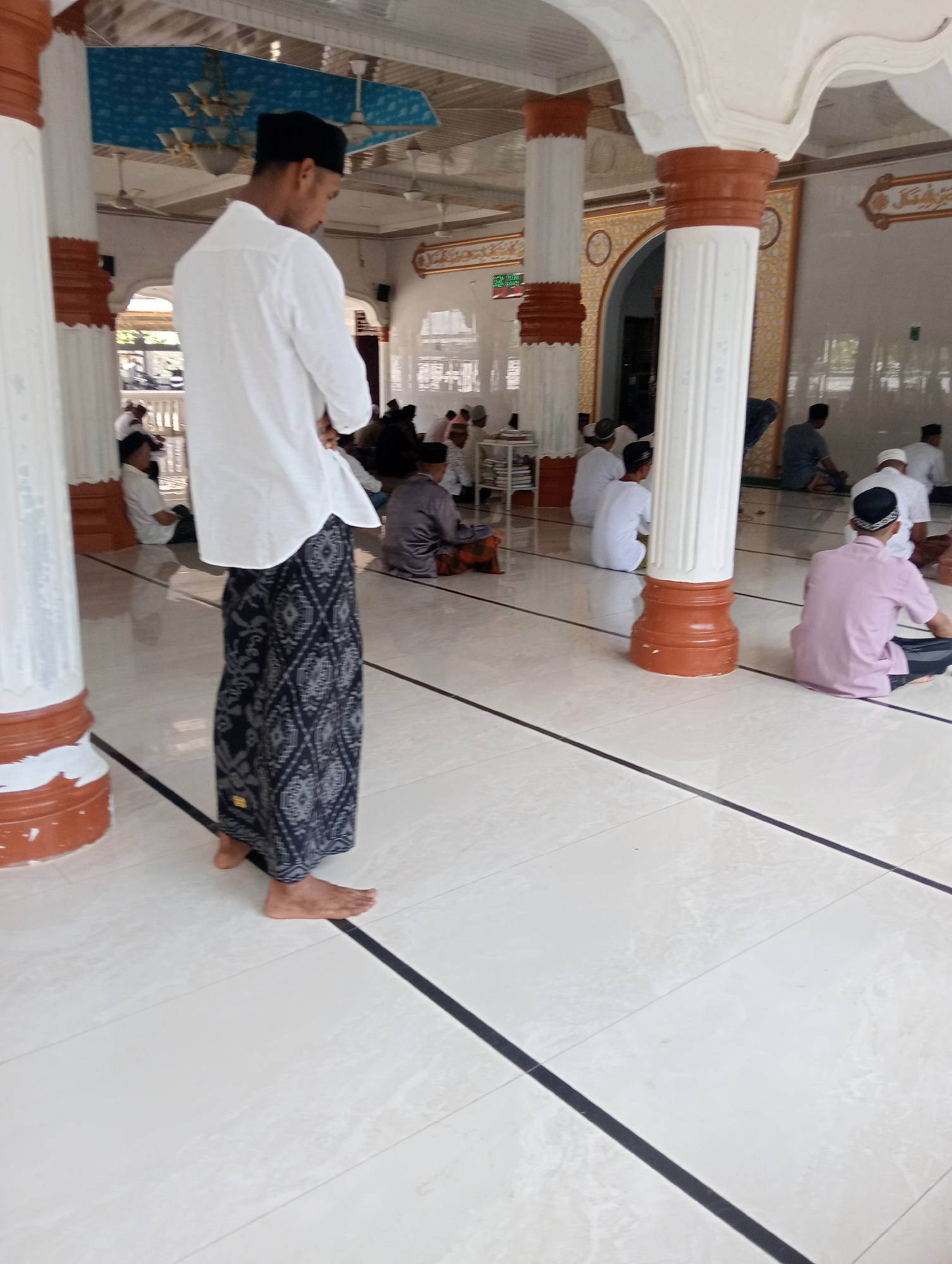 A full shot of a mosque's interior with people in prayer, taken in Lhokseumawe, Indonesia. A man in a black cap, white shirt, and sarong stands in the foreground, facing away from the camera. Several other individuals are seated on the floor in prayer. The mosque features white tile flooring with black lines, and white columns with brown accents. A chandelier hangs from the ceiling.