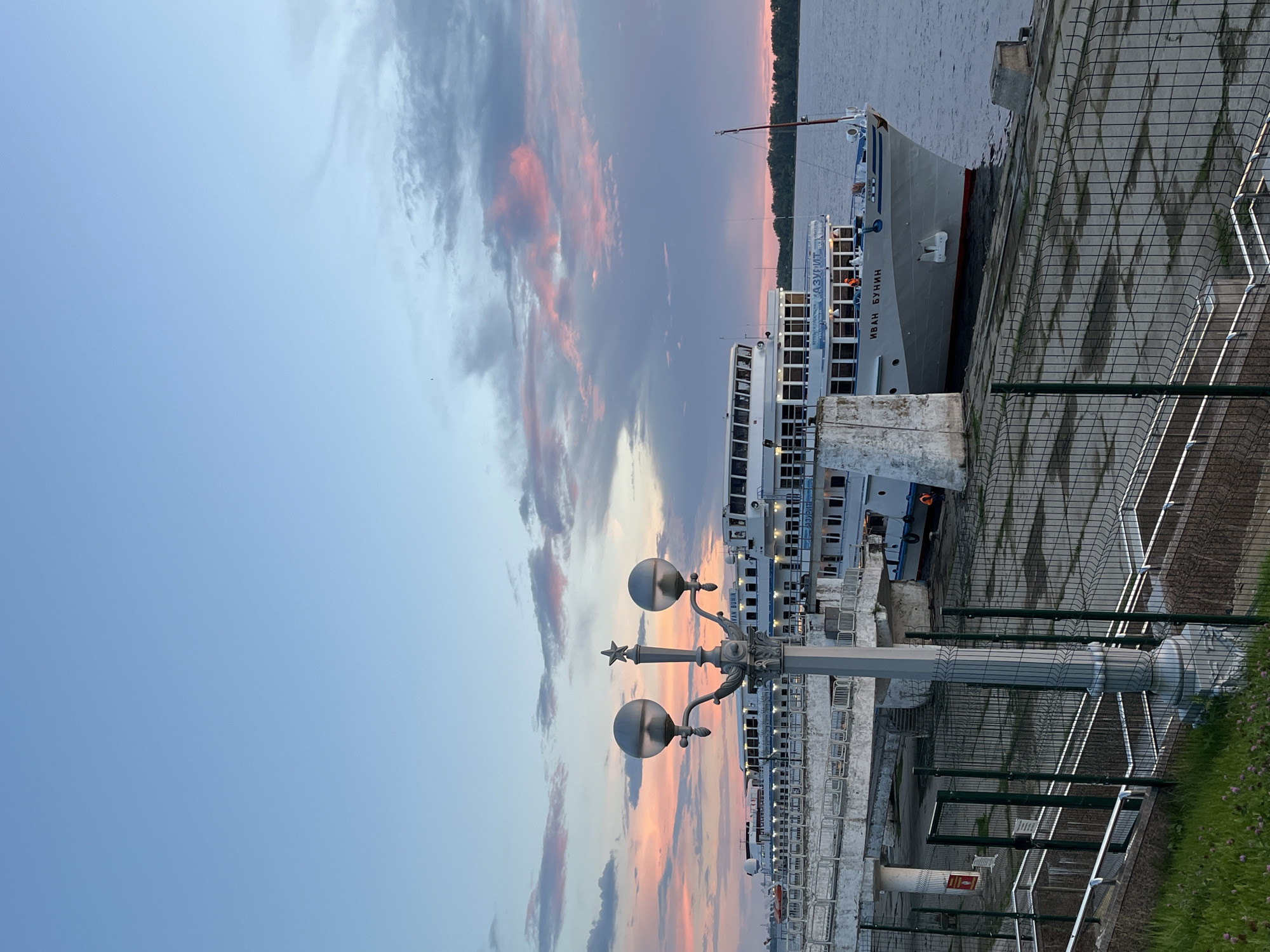 The image depicts a serene waterside scene at dusk in Novokuybyshevsk, Russia. The primary subject is a large white ship docked along a paved promenade. The ship, named "ИВАН БУНИН" (Ivan Bunin), is a passenger vessel, evident from its multiple decks and windows. There are no people clearly visible on the ship's exterior, though a figure in an orange vest can be seen on the deck.
The setting is a waterfront promenade with a concrete walkway and a metal fence bordering the edge. A decorative lamppost with two spherical lights and a star ornament stands prominently in the foreground, suggesting the onset of evening. The sky is a canvas of soft colors, with hues of pink, orange, and purple streaking through scattered clouds, indicative of a sunset. The water is calm, reflecting the sky's gentle tones. Beyond the ship and the water, a tree-lined horizon is faintly visible.
The overall mood is peaceful and tranquil, amplified by the soft light and the stillness of the water. The time of day is clearly dusk, transitioning into evening. The location is identified as Novokuybyshevsk, Russia, though specific landmarks are not apparent in this frame. The presence of the ship and the promenade suggests a port or a popular recreational area. The concrete surface, metal fencing, and the ship itself are key objects contributing to the urban waterfront atmosphere.