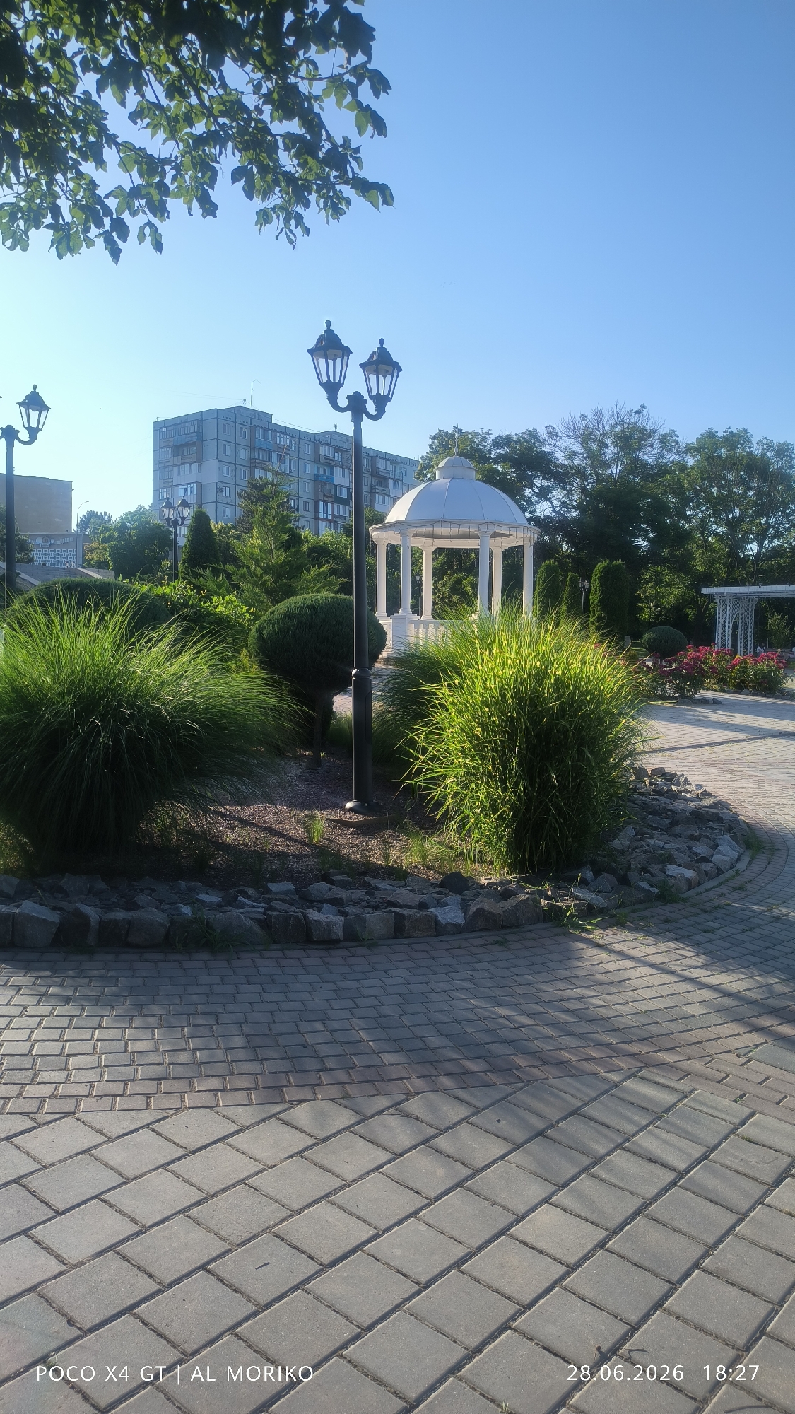 This media file is a vertical, eye-level, outdoor photograph of a park-like setting bathed in the warm glow of late afternoon sun. The scene is dominated by a white gazebo with ornate columns and a domed roof, suggesting it is a place for leisure or events. In the foreground, a paved pathway made of interlocking gray and reddish-brown bricks leads into the scene, with a noticeable shadow cast across it.
To the left of the pathway, a manicured garden bed features two large, fluffy ornamental grasses that catch the sunlight, along with a neatly trimmed, rounded shrub and other smaller greenery. A black lamppost with two ornate lanterns stands near these plants. Further into the background, a tall, multi-story building with numerous windows is visible, hinting at an urban or suburban environment surrounding the park. The sky is a clear, bright blue, indicative of fair weather.
The overall mood of the image is peaceful and serene, enhanced by the soft lighting and the well-maintained landscape. The presence of the gazebo and the paved walkways suggests a public space designed for relaxation and possibly gatherings.
Visible text at the bottom of the image reads: "POCO X4 GT | AL MORIKO 28.06.2026 18:27". This text indicates the device used to capture the photo, the photographer's name or watermark, and the date and time of the photograph.