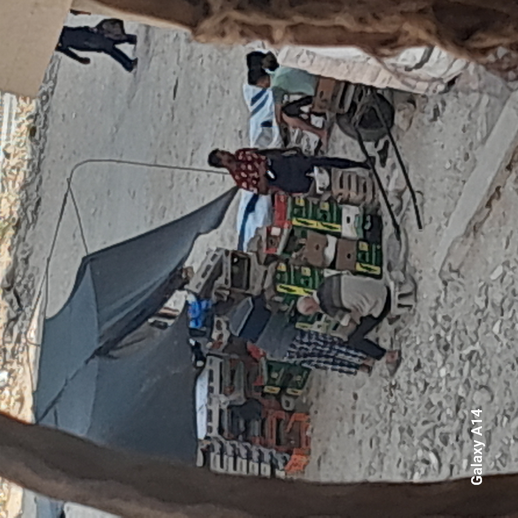 The image depicts an outdoor scene, likely a market or storage area, in Khan Yunis, Palestinian Territories. Several people are present. In the foreground, a person wearing a checkered shirt and dark pants is seated on a round object, possibly a stool, near stacks of boxes. To the right, another person in a red and white patterned shirt and dark pants stands holding a dark object. Further to the right, a person in a light blue shirt and dark shorts is seated on a crate. In the background, a person in dark clothing is walking away, carrying a dark bag. The environment consists of a sandy or dirt ground with numerous cardboard boxes and pallets stacked high, some of which appear to be filled with goods. A dark tarp is stretched over some of the structures. The overall impression is of a busy, albeit somewhat cluttered, commercial area.