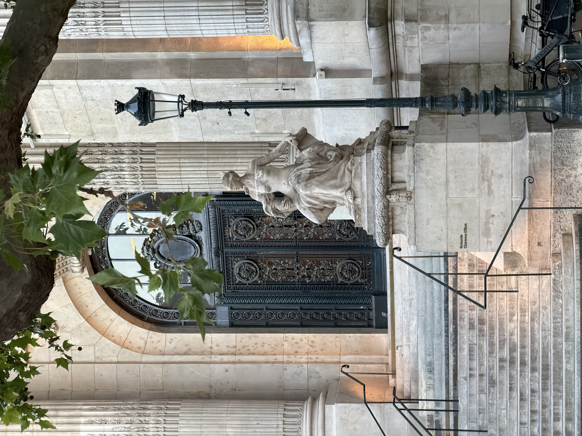 The image depicts the entrance to the Bibliothèque Nationale de France Richelieu in Paris. The architectural style features stone columns, a large arched doorway with intricate metalwork, and a stone statue of a reclining figure above the door. A vintage lamp post is situated near the entrance. A portion of a tree with green leaves is visible on the left side of the frame. A series of steps with metal railings lead up to the entrance. A bicycle is parked at the top of the stairs.