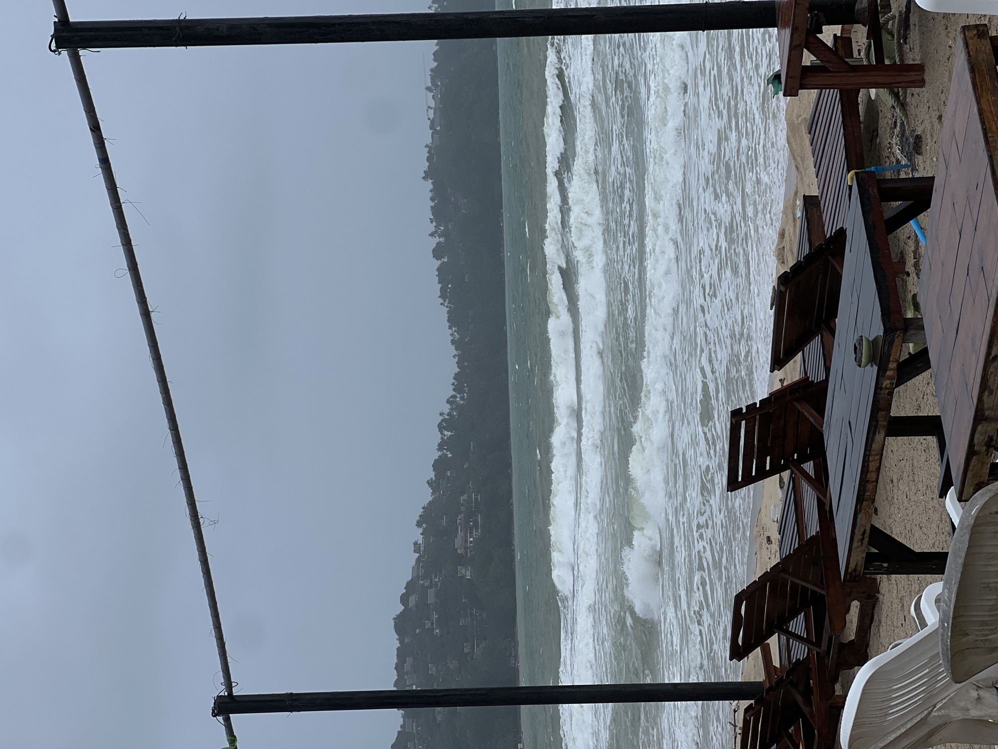 This media file captures a desolate beach scene under an overcast sky. The image is taken from a slightly elevated position, looking out towards the ocean.
In the foreground, elements of a structure are visible: two dark, horizontal beams, likely part of a canopy or railing. A thinner, lighter-colored rod or wire is stretched across the frame, secured to the beams with several small ties. To the right, on what appears to be a sandy beach, several wooden picnic-style tables and benches are present, along with a stack of white plastic chairs. The wooden furniture appears wet, suggesting recent rain.
The midground is dominated by the ocean, characterized by significant, white-capped waves crashing towards the shore, indicating rough sea conditions. Beyond the turbulent water, a lush, dark green hillside rises in the background, densely covered with trees. Scattered buildings or houses can be faintly seen nestled among the foliage on the hillside.
The sky is a uniform, dull grey, suggesting heavily overcast weather. There are no people visible in the scene, and the overall atmosphere is one of quiet solitude, possibly due to the inclement weather. The time of day is not explicitly clear, but the diffused light is consistent with an overcast day. There is no visible text in the image.