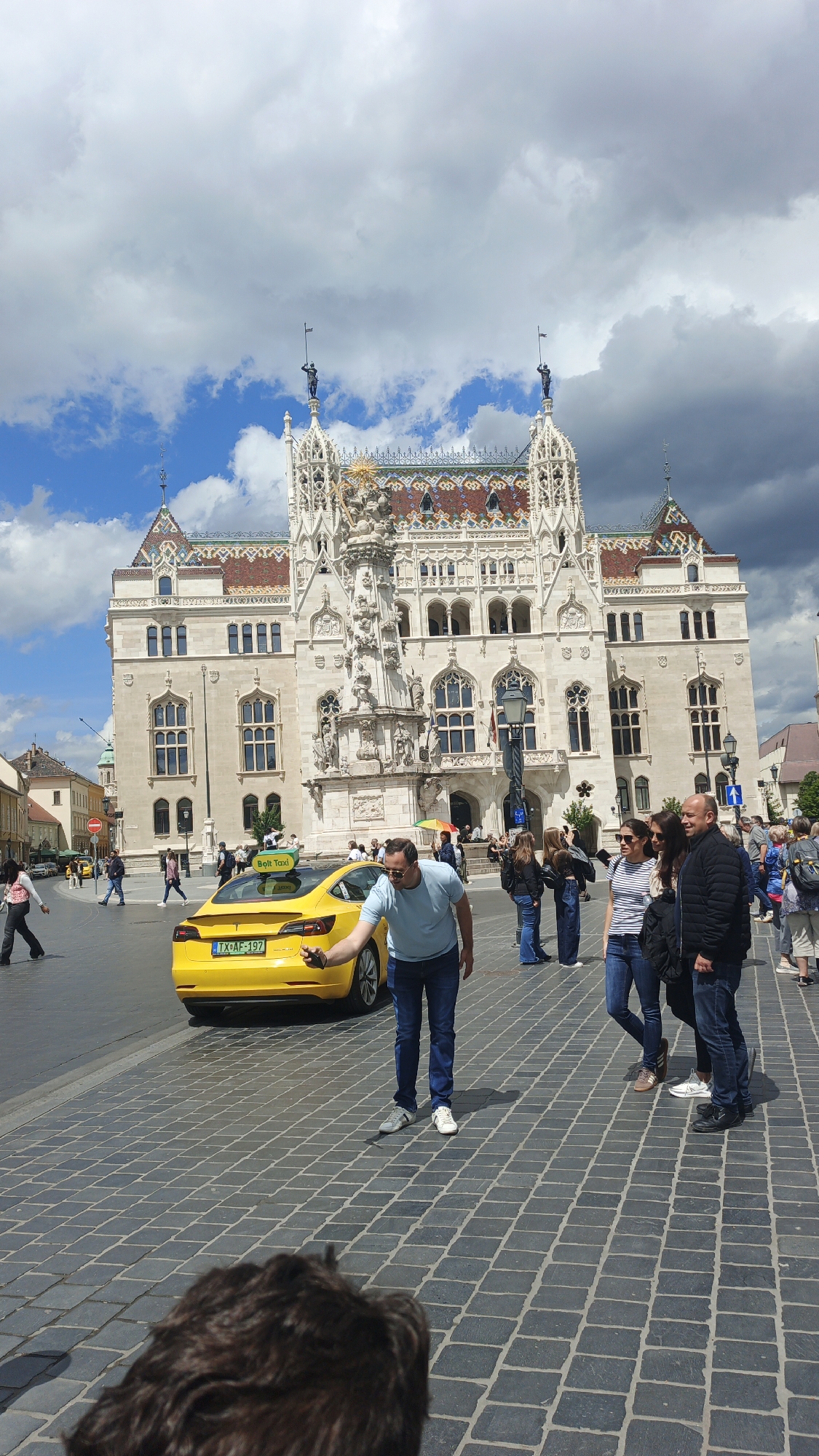 The image shows a bustling outdoor scene in Budapest, Hungary, on a partly cloudy day. A yellow Tesla taxi with "Bolt Taxi" signage on its roof is parked near a crosswalk. A man in a light blue shirt and jeans is standing next to the taxi, looking at his phone and gesturing with his right hand. Several other people are visible in the background, including a group of three people standing close together, and other pedestrians walking in the distance. The backdrop is a large, ornate building with multiple windows and decorative elements, likely a historic landmark. The ground is paved with dark grey rectangular stones.
