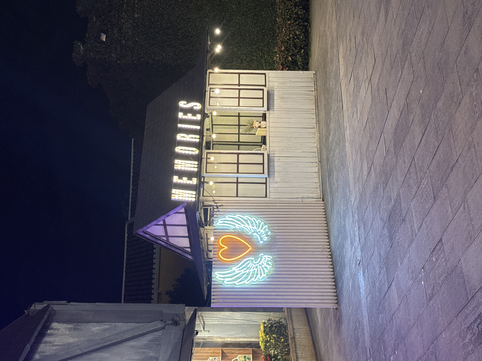 The photograph depicts a nighttime scene in Hội An Ward, Vietnam, showcasing a building exterior. The structure is adorned with the illuminated word "MEMORIES" vertically on its facade. Below this, a neon sign in the shape of a heart with wings is visible. The building has an open section with windows, hinting at interior space. The surrounding environment includes greenery and paved ground.