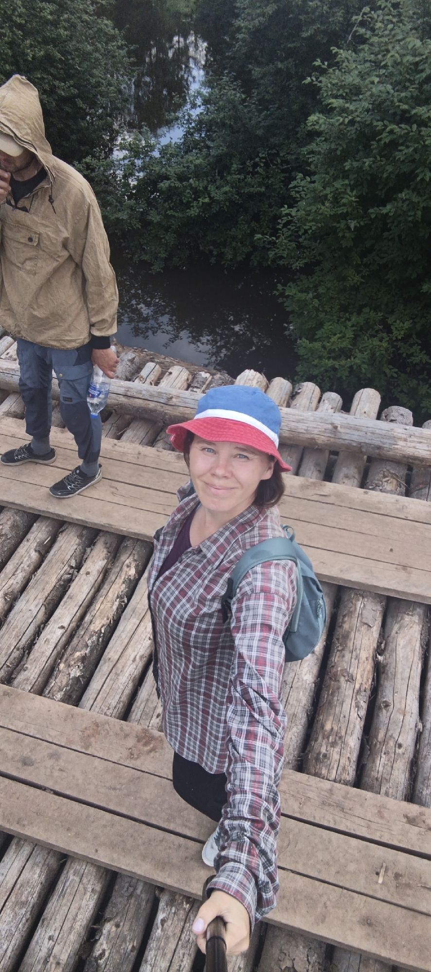 A medium-shot, high-angle selfie shows two individuals on a rustic wooden bridge over a body of water. In the foreground, a person wearing a red and blue bucket hat, a plaid shirt, and a backpack smiles at the camera, holding a selfie stick. To the left, further back on the bridge, another person in a hooded tan jacket and blue pants holds a clear plastic water bottle. The bridge is constructed from thick logs laid horizontally and supported by vertical wooden posts. The background features dense green foliage on either side of the water, which appears dark and still. The scene is set in a natural environment, possibly in or near the city of Garevaya, Russia, given the context.