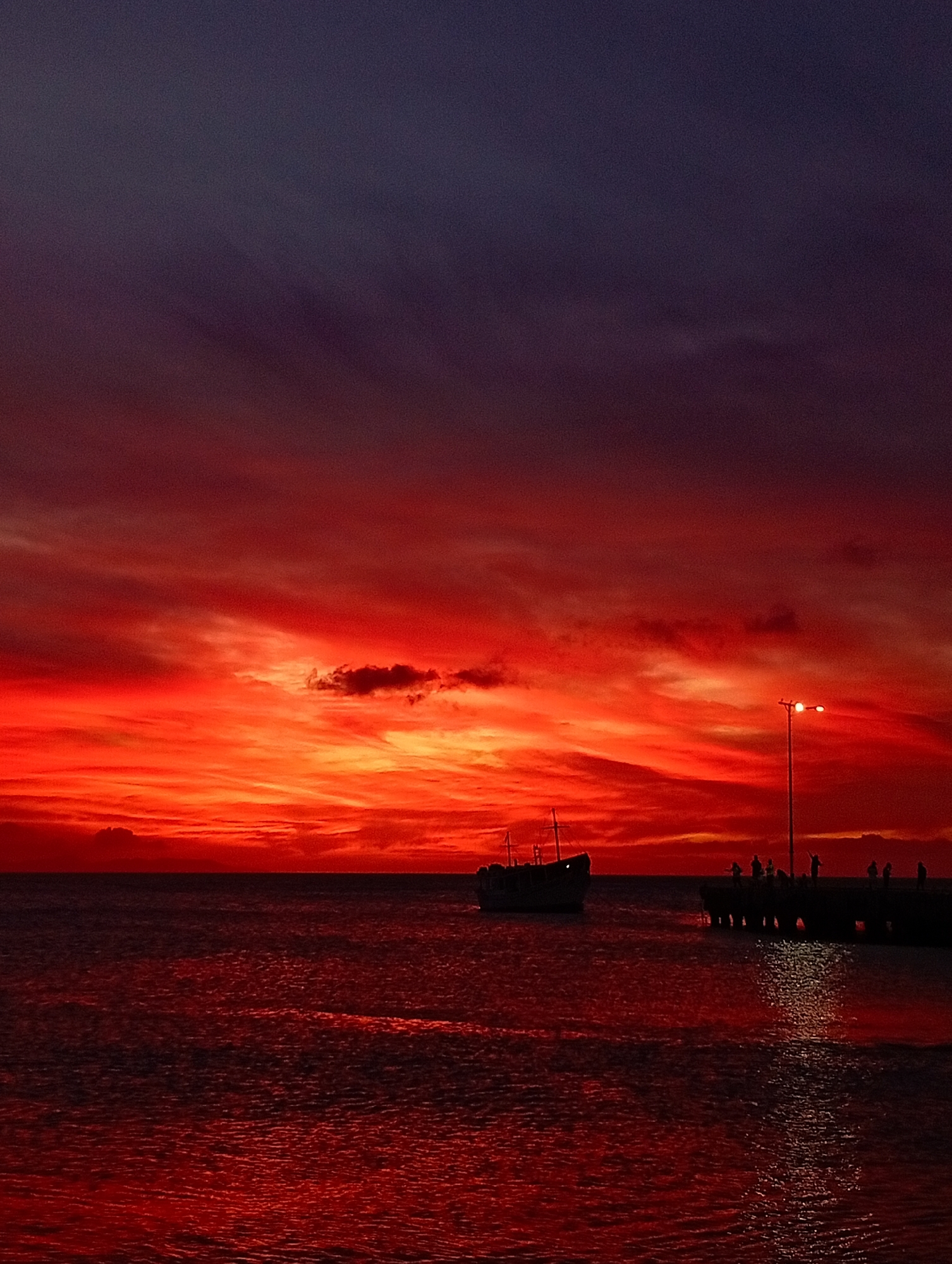 The image displays a dusk scene in Juan Griego, Venezuela. The sky transitions from a deep purple-blue at the top to a vibrant, intense red and orange across the middle and lower sections, featuring discernible cloud streaks. Below this horizon, a body of water, likely the sea, reflects the sky's red tones, appearing dark red with visible ripples. In the mid-ground, a dark-silhouetted boat with a mast is positioned on the left side of the frame, partially submerged in the water. To the right, a dark, elongated pier extends into the water. Multiple dark-silhouetted human figures are observed standing on the pier. A single streetlamp, illuminated and equipped with two lights, stands on the pier, casting a prominent vertical reflection on the water's surface. No specific actions or interactions among the individuals are clearly discernible due to their silhouetted forms against the bright sky.