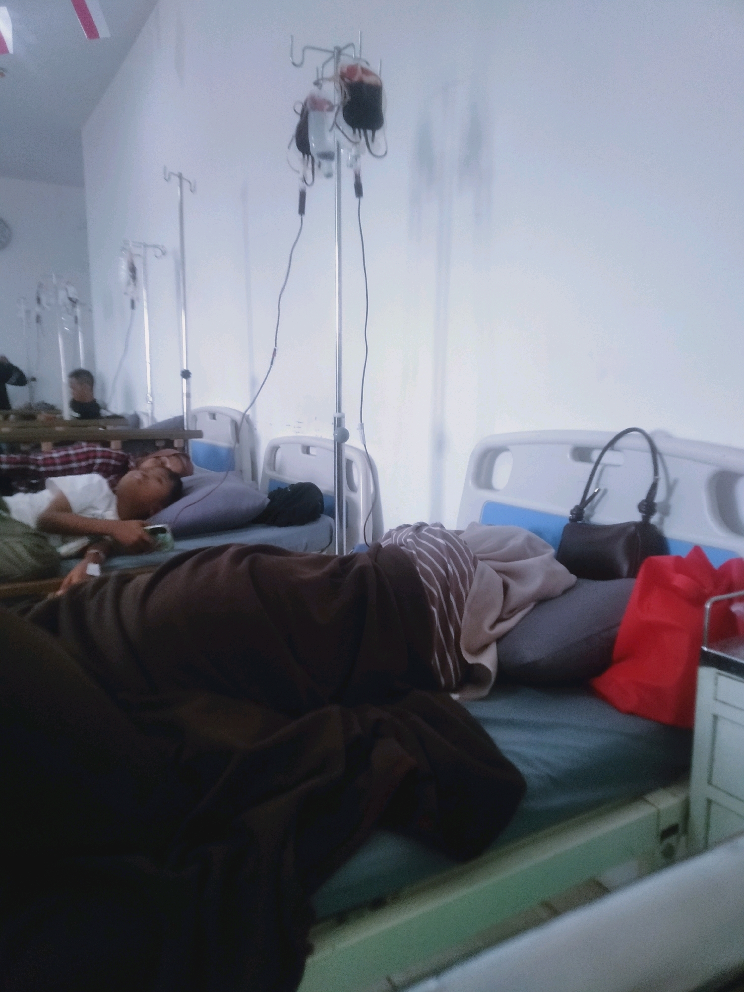 The image depicts a hospital ward in Cianjur, Indonesia, with multiple patients in beds. In the foreground, a person lies in bed covered by a dark blanket, their arm resting on a pillow. Beside them, another patient is seen from the shoulders up, looking towards the viewer. Further back, other patients and hospital beds are visible, along with IV stands and bags of fluid. The room appears to be a shared space, with medical equipment and personal belongings scattered around. The overall impression is one of a busy and functional healthcare setting.