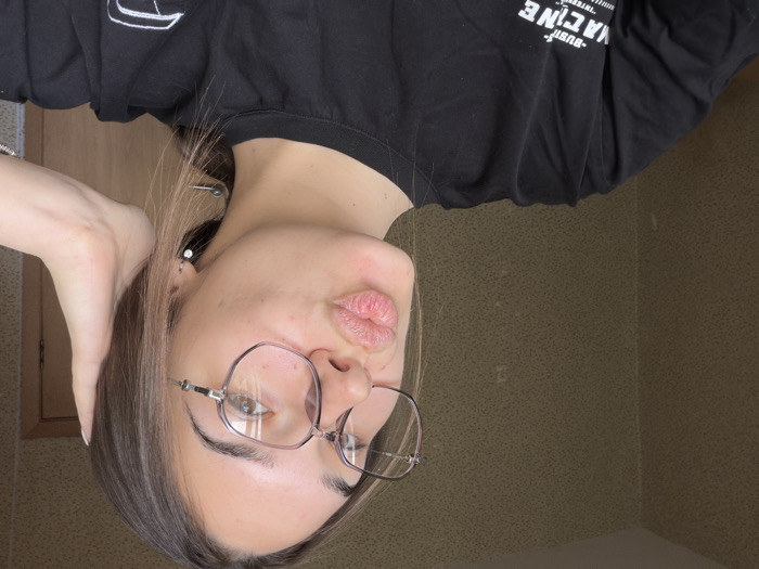 An indoor scene, likely in Sofia, Bulgaria, features a young adult female captured in a close-up, self-portrait style photograph. The subject, positioned centrally, has brown hair, wears light-colored, possibly clear, octagonal-framed prescription glasses, and has her lips pursed in a "kiss" expression. Her left hand is partially visible, with fingers touching the back of her head on the right side of the frame. She wears a small, silver-colored earring with a heart-shaped charm in her visible left ear. Her attire consists of a black short-sleeved t-shirt displaying partially visible white text, including "BUSH-" and "MACINE". The background reveals a textured, light brown wallpapered wall on the left and a portion of a wooden door and door frame on the right. The lighting appears to be ambient, illuminating the subject from the front.