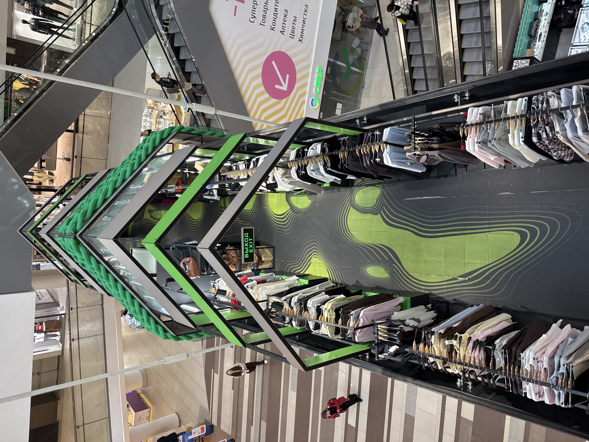 This image shows the interior of a shopping mall, likely located in Saint Petersburg, Russia, based on the Cyrillic text on signage. The camera is positioned high, looking downward at an angular clothing rack display in the foreground. The clothing rack is made of black metal frames with green accents, and it has several tiers of clothes hanging on racks. A large green rope-like design runs along the top of the rack.
The floor is decorated with abstract green wave patterns, with portions of the floor consisting of light and dark brown stripes. People can be seen walking around the display.
In the background, escalators are visible, along with signs in Russian indicating the types of stores available in the mall (e.g., "Supermarkets," "Candy," "Pharmacy," "Flowers," "Dry Cleaning"). There are other levels and shops visible in the distance, as well as people on the escalators and in the surrounding area of the mall.
