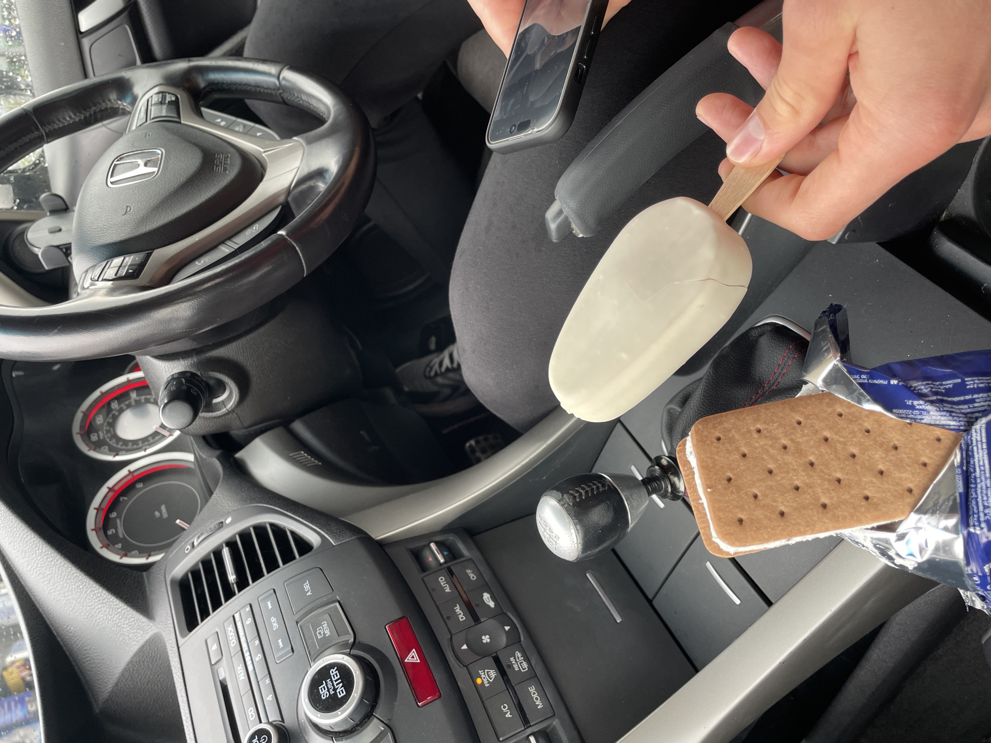 The image captures the interior of a car during what appears to be an overcast day. A person is visible from the waist up, holding a white chocolate-covered ice cream bar on a wooden stick in their right hand. Their left hand holds a smartphone.
The car's interior features a black Honda steering wheel with controls on both sides and a silver Honda logo. The dashboard includes an analog speedometer and tachometer, air vents, and an audio/climate control panel. Visible buttons and labels on the control panel include "A/C," "MODE," "FRONT," "REAR," "AUTO," "OFF," "DUAL," "AUX," "CLOCK," "CD," "SKIP," "MENU," "SEL," "PUSH ENTER," "A.SEL," and a red hazard warning button. A manual gear shifter with a black knob is positioned in the center console.
Next to the gear shifter, an open package of an ice cream sandwich is resting. The sandwich features two rectangular, brown, perforated cookies with a white filling in between. The wrapper, partially visible, contains text in Hebrew and English, including "חול טל מסחר והשקעת AR" and "Palestine EL:02-2220056", along with "αριθ. 21," and "9600222-20". The driver's seat and floor mat are also visible. The overall scene suggests a break or snack time inside the vehicle.