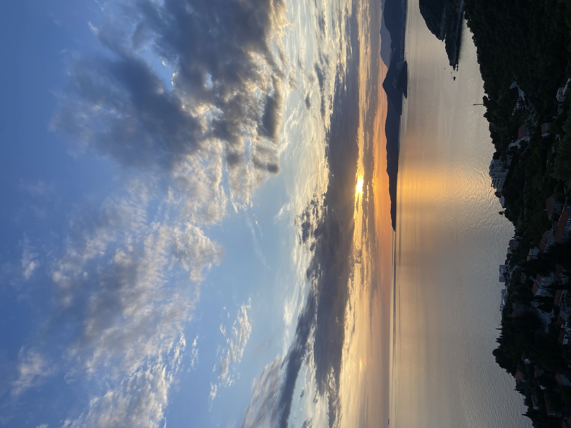 This image captures an expansive coastal landscape at sunset, located in the city of Unknown, Unknown, viewed from a high vantage point overlooking the sea. The sky transitions from a deep clear blue at the top to vibrant orange and yellow hues near the horizon. Numerous clouds, varying from wispy cirrus to darker, more dense stratocumulus formations, traverse the sky, with some obscuring the sun. The sun, a brilliant orange orb, is partially hidden by a dark cloud band directly on the horizon, casting an intense golden path of light across the calm sea surface.
A vast body of water, likely a sea or ocean, occupies the middle ground, reflecting the sky's colors in varying shades of gold, orange, and muted grey. On the far left horizon, a faint, dark, rectangular vessel is visible on the water. The foreground features a verdant coastline with steep, tree-covered slopes. Scattered residential buildings with red-tiled roofs are nestled among the dark green trees on the lower left hillside. A prominent, multi-story, light-colored building complex with architectural details stands on a promontory extending into the water on the mid-right, surrounded by lush foliage. Winding roads or paths are visible traversing the landscape. A slender, dark tower-like structure is also visible near the larger building. In the background, a series of dark, undulating mountain ranges and hills recede into the distance, silhouetted against the lighter horizon. No individuals or discernible human activity are present within the frame.